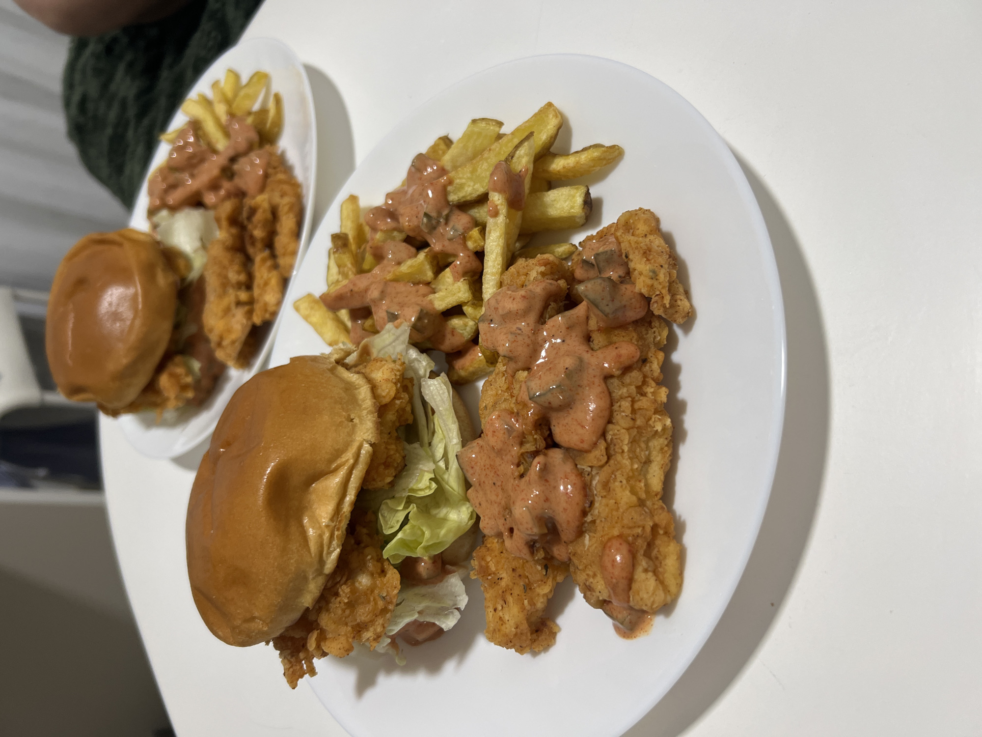 The image shows two white plates of food. Each plate contains french fries, fried chicken, and a sauce, possibly fry sauce, on top. One plate also has a sandwich on it. The scene appears to be indoors, on a white surface. The location in the city of Mogán, Spain is not identifiable from the image.