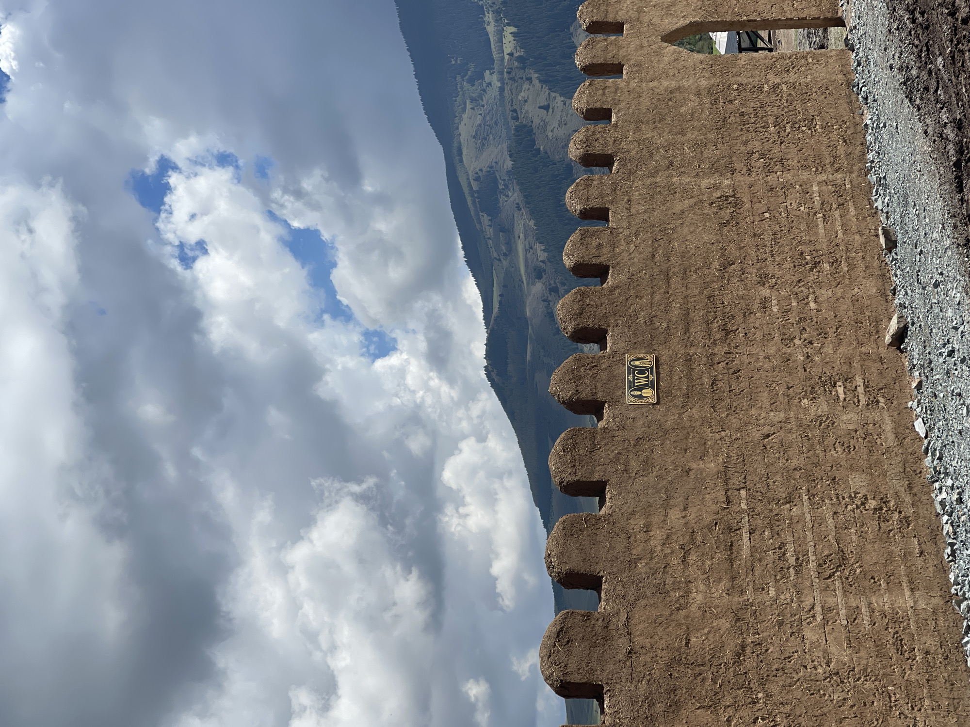 A close-up, low-angle shot depicts a crenellated mud-brick wall in Semenovka, Kyrgyzstan, against a dramatic sky filled with heavy, grey clouds and patches of blue. The wall, constructed from rough-hewn earth, has a textured, uneven surface, with horizontal lines of embedded straw or wood visible. The battlements at the top are rounded, resembling teeth. A small, ornate sign with the letters "WC" and silhouettes of a male and female figure is affixed to the wall. To the right, an arched opening reveals a glimpse of a structure beyond. The foreground is a mix of gravel and dark, damp soil, suggesting a construction site or recent rain. In the background, rolling, tree-covered hills rise towards the horizon under the ominous sky. No people are visible in the frame.