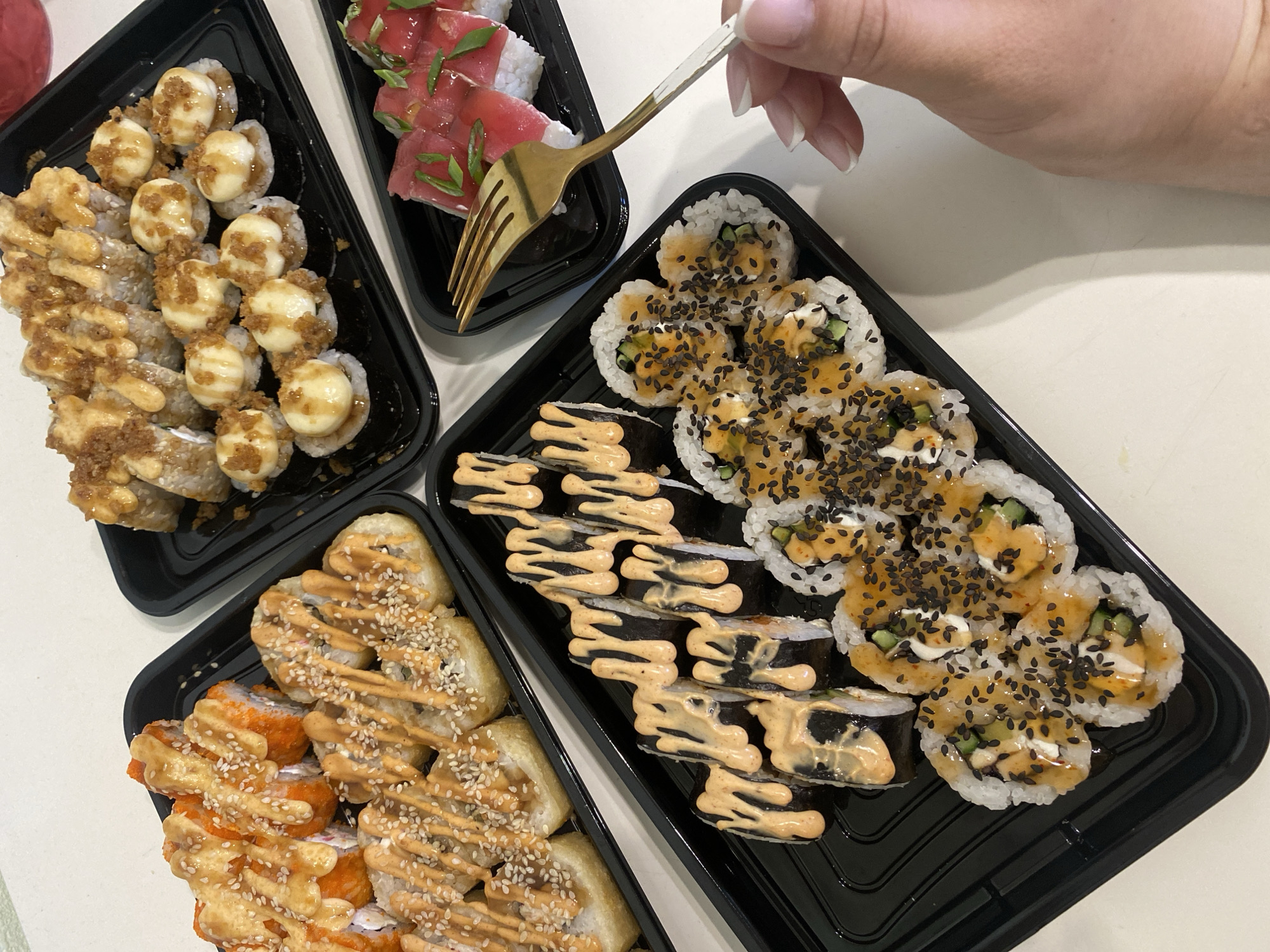 The image captures an overhead view of a diverse selection of sushi rolls arranged in four black plastic takeout trays on a light-colored surface, likely a tabletop. A person's right hand is visible in the upper right quadrant, holding a golden-colored fork with a clear handle, poised over one of the sushi trays, suggesting the act of eating or serving. The hand has light-colored nail polish.
The sushi trays feature a variety of rolls, each with distinct toppings and presentations. In the top left tray, rolls are generously topped with creamy white dollops and a brownish, crispy garnish, possibly fried onions or tempura flakes, along with a light drizzle. The top right tray contains what appear to be inside-out rolls or nigiri, topped with slices of red fish, likely tuna or salmon, and garnished with finely chopped green herbs.
The bottom left tray holds two types of rolls: one with an outer layer of orange roe, drizzled with a light orange sauce and sprinkled with sesame seeds, and another set of rolls also adorned with sauce and sesame. The bottom right tray showcases maki rolls wrapped in dark nori, artfully drizzled with a zigzag pattern of light orange sauce, alongside inside-out rolls topped with a lighter orange sauce, black sesame seeds, and visible pieces of cucumber and a white filling.
The overall setting is informal, indicative of a takeout meal or a casual dining experience. The lighting is bright and even, consistent with an indoor environment during the day or well-lit artificial light. No text is visible in the image. The presence of multiple sushi varieties suggests a shared meal or a large order, originating from a location in Sochi, Russia.
