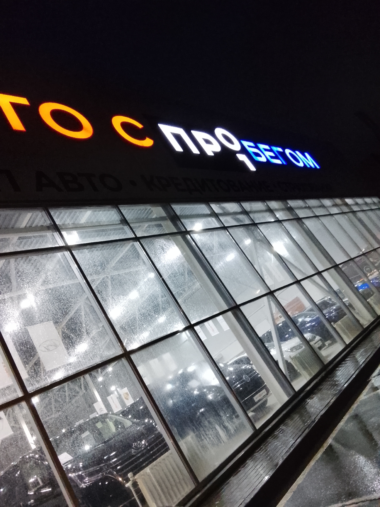 The image depicts the exterior of a car dealership in Obninsk, Russia at night. The building has a large glass facade with multiple panes reflecting the interior lights and parked cars. The signage "ГОС ПРОБЕГОМ" is illuminated in orange, white, and blue letters above the entrance. "ГОС" is lit in orange, "ПРО" in white and "БЕГОМ" in blue. The windows are wet, suggesting recent rain. Cars are visible inside the dealership, including a black sedan. The scene is angled slightly upward toward the building.
