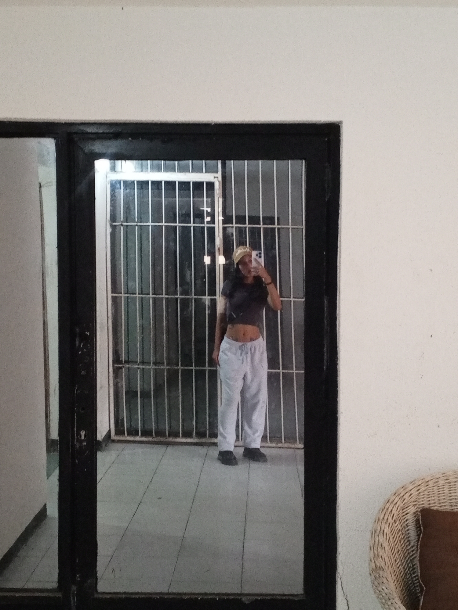 A woman is taking a selfie in a mirror. She is wearing a crop top, sweatpants, and a baseball cap. Behind her is a barred window. The floor is tiled. To the right of the mirror is a white wall and the edge of a wicker chair. The location may be in Porlamar, Venezuela.