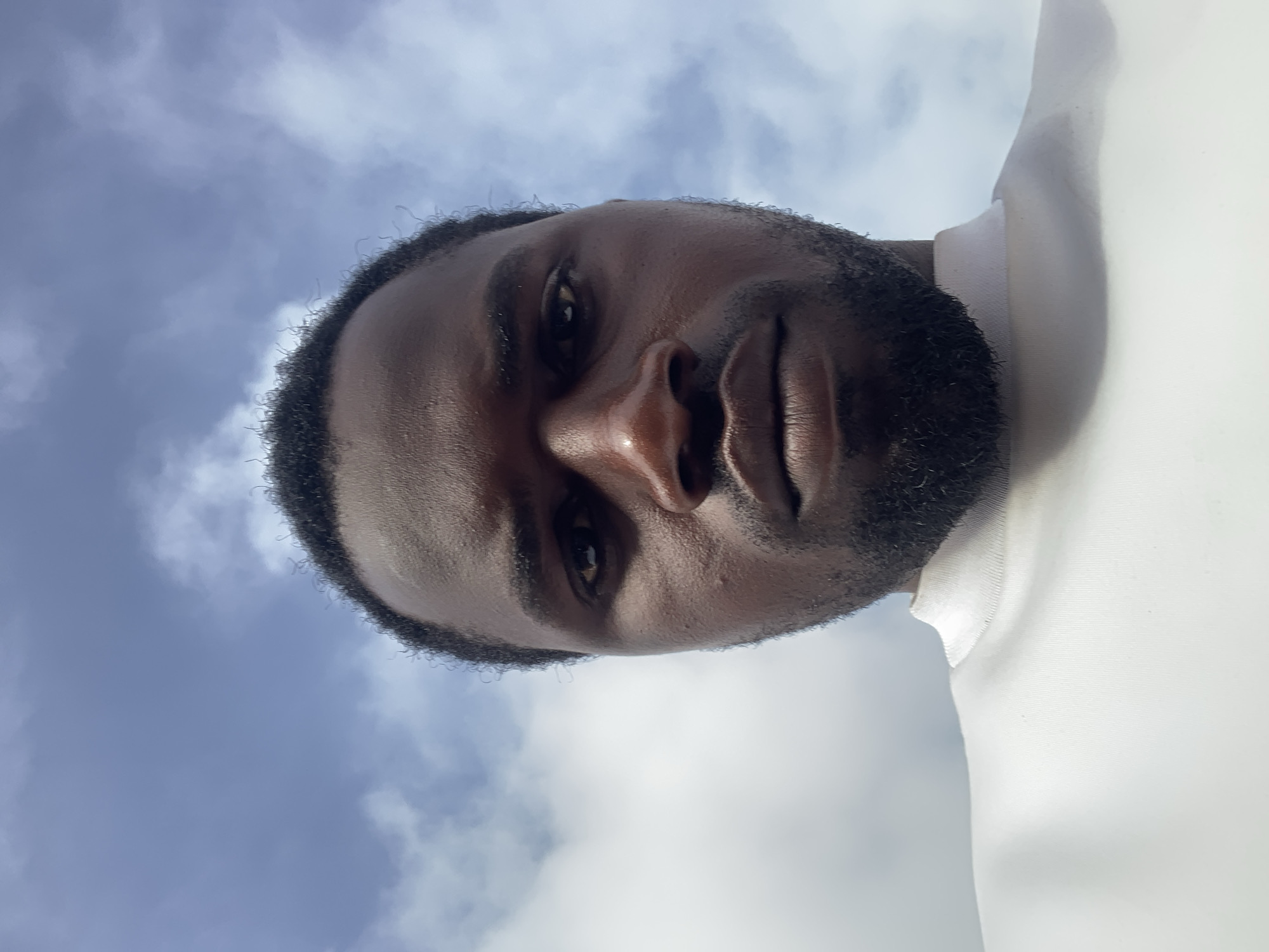 An outdoor portrait captures a man with dark skin and a short beard, looking directly at the camera from a low angle. The image is rotated, presenting the man's face diagonally across the frame. He has dark hair, and his facial features are well-defined, with a serious or neutral expression. He appears to be wearing a light-colored, possibly white, collared shirt, visible on the right side of the frame.
The background consists of a bright blue sky with scattered white clouds, indicating it is daytime. The lighting suggests natural sunlight. There are no specific objects or activities visible beyond the man and the sky. No text is visible in the image. The scene is set in what appears to be Barakin Lerawi, Nigeria.