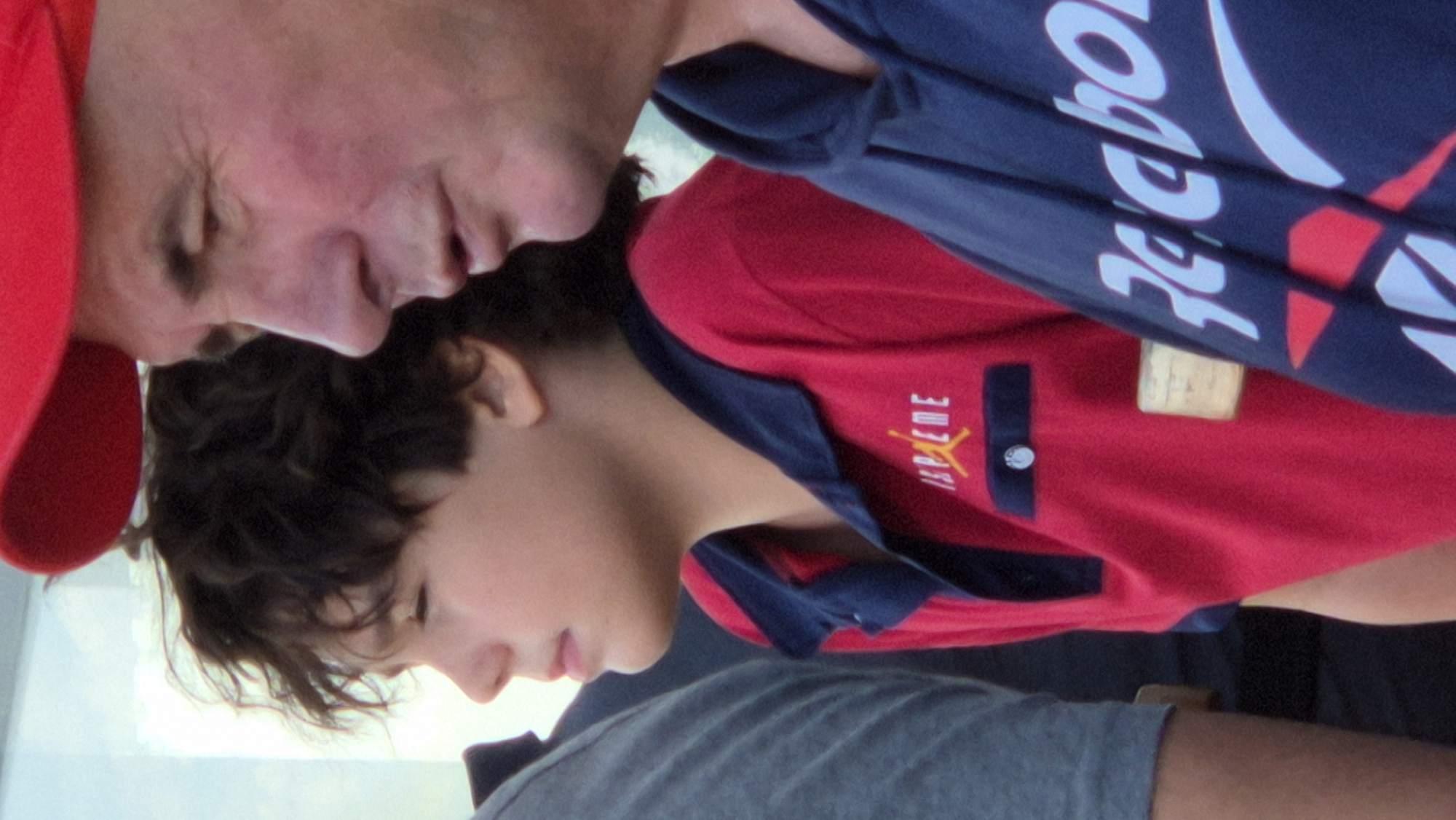 The image shows a close-up of three people from the shoulders up, in an outdoor setting that appears to be a public space in Promyshlennaya, Russia. In the foreground, on the right, a man wearing a red baseball cap and a blue hoodie is partially visible, his face turned to the side. To his left, a boy with dark, curly hair is wearing a red polo shirt with a blue collar and a logo that appears to be a stylized "MJ" with wings, possibly indicating Michael Jordan. His expression is neutral as he looks to his left. To the far left, another person's shoulder and arm are visible, wearing a grey t-shirt. The background is blurred and out of focus, suggesting a shallow depth of field, with elements that could be part of an urban environment or a park. The lighting is natural, indicating it is daytime.
