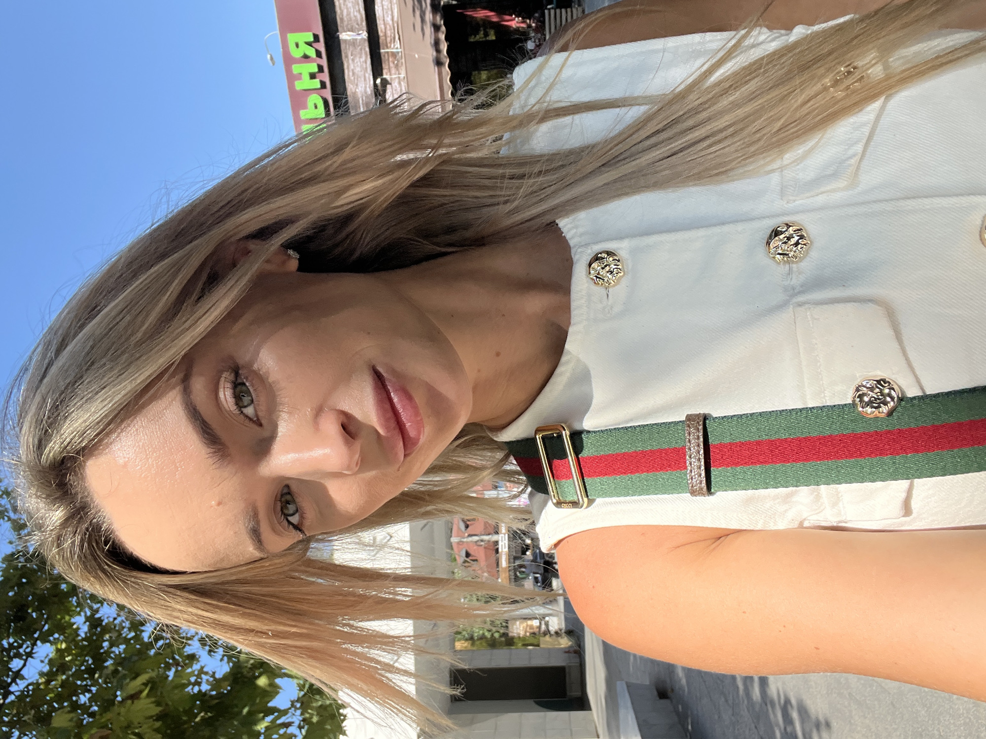 A blonde woman with green eyes, wearing a white vest and a striped Gucci strap, takes a selfie outdoors on a sunny day in Odesa, Ukraine. The background shows a building with green lettering that reads "ВЕРХНЯ", indicating a shop name. Trees and a blue sky are also visible.
