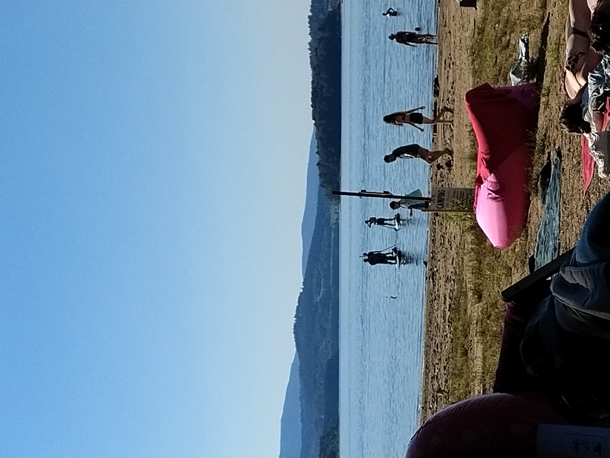 In the city of Radslav, Czechia, on a sunny day, people are enjoying a body of water. In the foreground, a person is relaxing on the beach on a pink beanbag chair, and another person is lying on a blanket. Further back, several individuals are paddleboarding on the lake. In the background, a lush green forested hill rises against a clear blue sky.