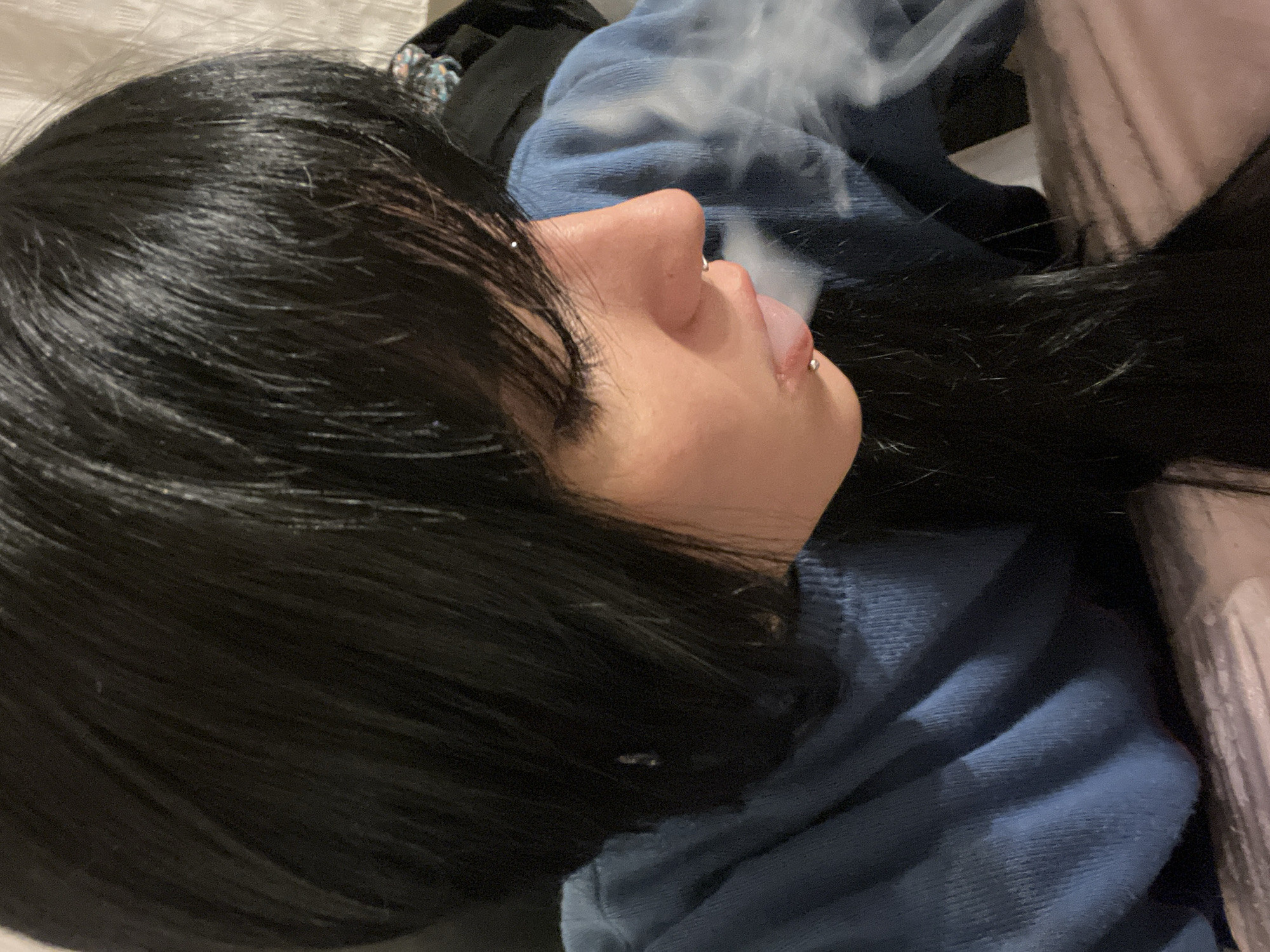 The image is a close-up, vertically oriented (though presented here rotated counter-clockwise), focusing on the left side profile of a person's head and upper torso. The person has long, dark, straight hair that appears sleek and somewhat wet-looking, covering part of their face and falling over their shoulders.
They are wearing a blue, ribbed or textured, long-sleeved garment, likely a sweater or hoodie. A small silver stud or ring piercing is visible in their left nostril, and a silver barbell piercing is seen on their slightly extended tongue. A prominent cloud of white vapor or smoke is emanating from their mouth, indicating they are exhaling from vaping or smoking.
The background is softly focused and indistinct, showing various folded fabrics in dark tones. On the far right, a light-colored, possibly beige or pinkish, ribbed fabric or bedding is visible. A multicolored hair tie or accessory is faintly discernible among dark fabrics behind the person's head. The scene suggests an informal indoor setting, possibly a casual space within a home in Buenos Aires, Argentina. The lighting is artificial, and no specific time of day can be determined. There is no visible text in the image.