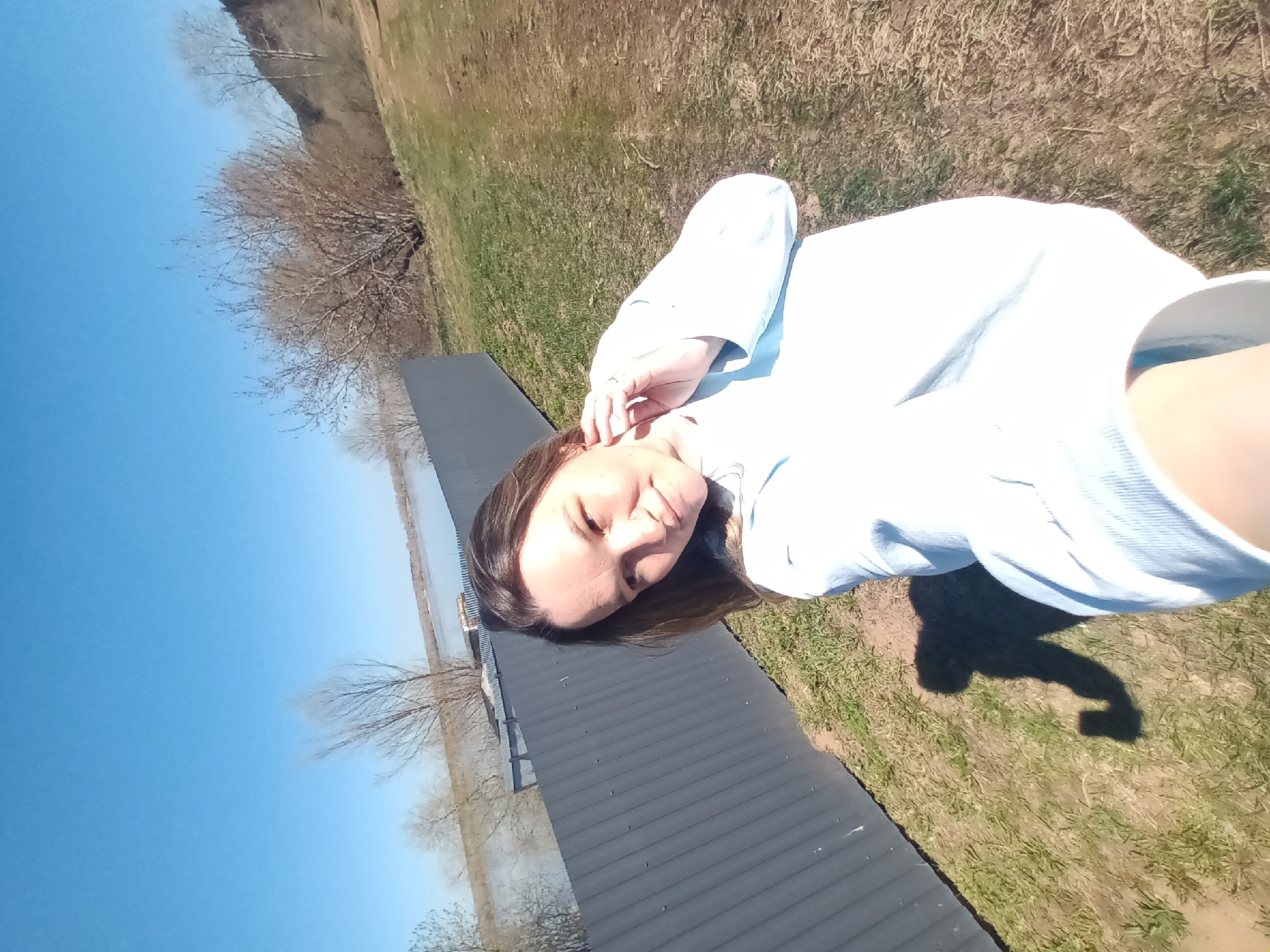 A woman lies on a grey, corrugated metal surface. Her head is near the bottom edge of the surface, and her torso and arm stretch towards the top. She wears a light blue shirt. Her shadow is visible on the grass to the right.
Behind the metal surface is a body of water and bare trees. The sky is blue. Beyond the grass on the upper right is a hill with more bare trees.
The location is Ольховка, Russia.
