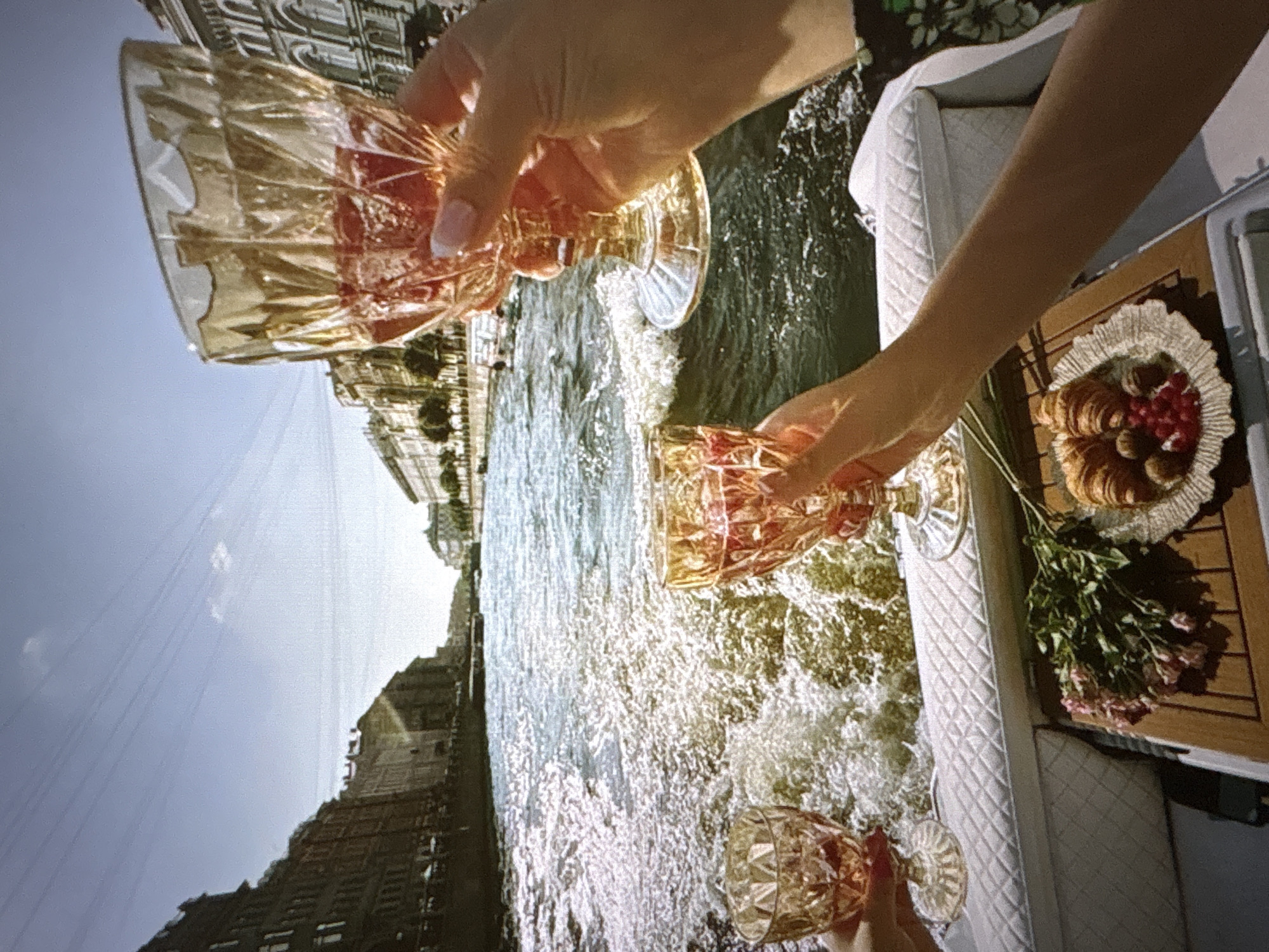 The image captures a celebratory scene on a boat on a river or canal, with buildings lining the banks. Two individuals are holding ornate glasses filled with a pinkish beverage, seemingly toasting. A small platter on a wooden table holds croissants, red berries, and chocolates. In the background, the water is disturbed, indicating the boat is in motion. The architecture suggests an urban setting, possibly European. The mention of Pori, Finland, places this scene within that specific city, suggesting the river or canal is likely one of Pori's waterways.