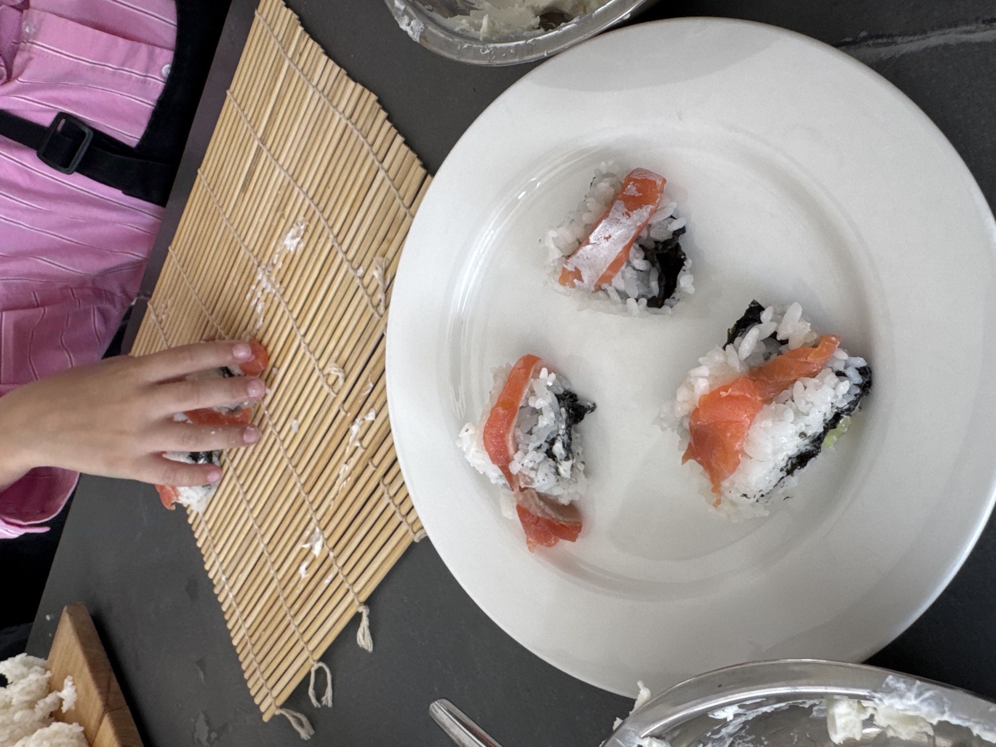 The image shows a close-up, overhead view of a person's hands making sushi. The hands are in the process of arranging slices of salmon on top of sushi rice, which is rolled in nori (seaweed) and placed on a bamboo rolling mat. There are three partially made sushi rolls on a white plate in the foreground. In the background, a wooden cutting board with a pile of white rice is visible. The scene appears to be indoors, likely in a kitchen. There is no information in the image to suggest it is located in Izhevsk, Russia.