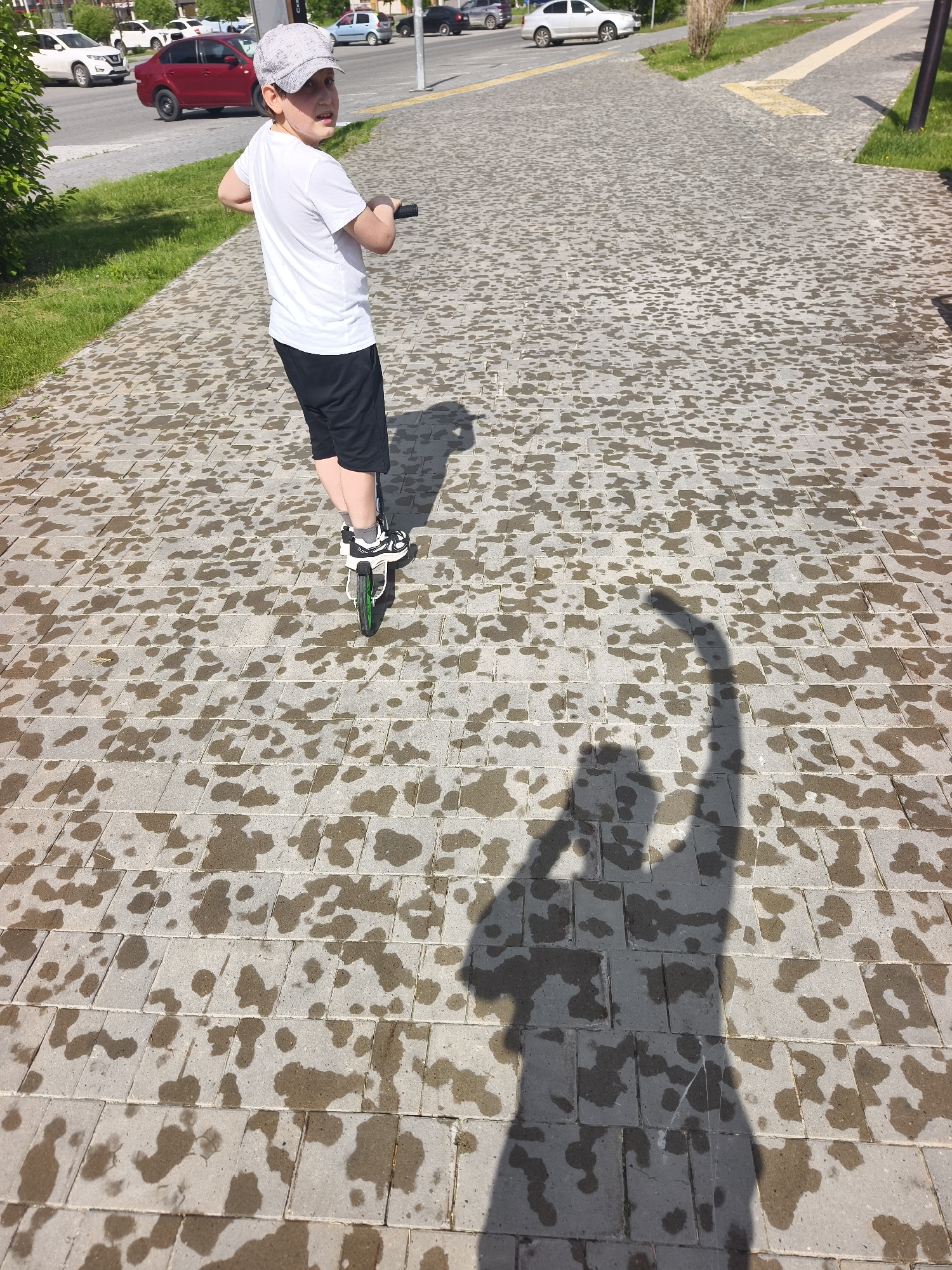 A child wearing a grey cap, white t-shirt, and black shorts is standing on a scooter on a paved surface. The pavement is wet with a pattern of darker patches, suggesting recent rain or cleaning. The child is looking over their shoulder towards the left side of the frame. A dark shadow, likely of the person taking the photo, is cast on the pavement, with one arm raised. In the background, there are several parked cars and some greenery. The location is identified as Tyumen, Russia.