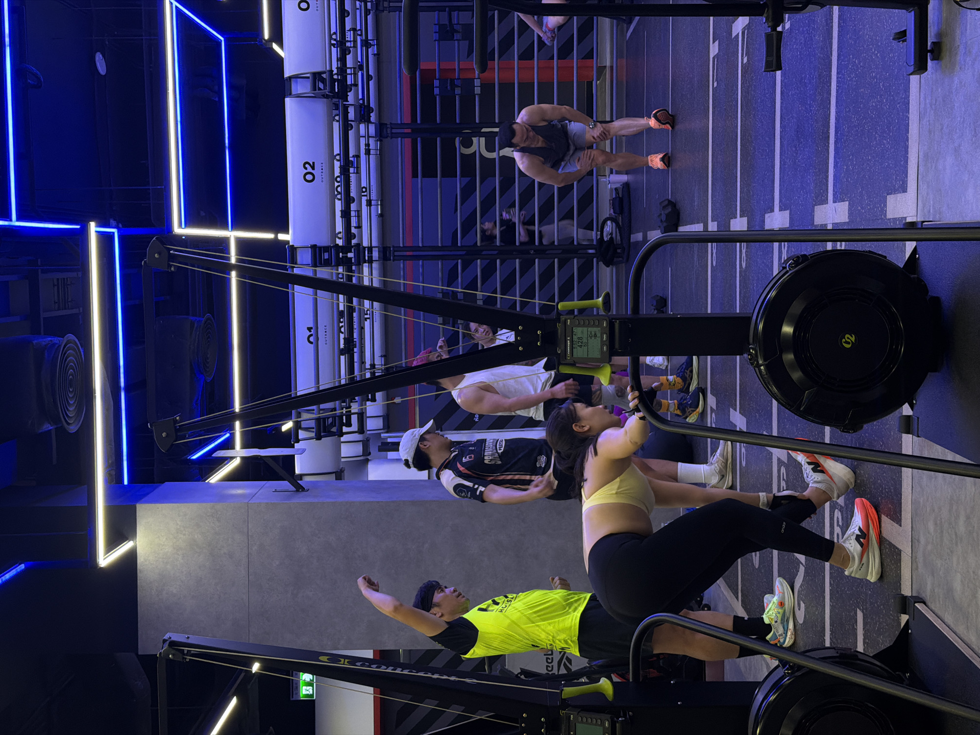 This media file depicts a group of people engaged in a workout at a gym. The scene is characterized by bright blue and yellow neon lighting, creating a modern and energetic atmosphere. The primary objects in the foreground are rowing machines, with several individuals actively using them.
In the middle ground, more people are visible, some on rowing machines and others standing or sitting. The background shows gym equipment, including weight racks and bars, as well as large white cylindrical structures with "OUTRACE" and numbers like "02" and "15" printed on them, suggesting a branded fitness facility. The floor is marked with white lines, likely indicating workout stations or lanes.
The individuals are dressed in athletic wear, indicating they are participating in a fitness class or training session. One person in the foreground wears a bright yellow shirt and black pants, while another wears a white tank top and black shorts. The overall mood appears to be focused and active, consistent with a gym environment. The visible text includes "OkConcer," "eel," "HUISL," "PRAYA," and numerical data on the rowing machine consoles, as well as branding on the cylindrical structures. Given the context of Nong Pa Khrang, Thailand, it is likely daytime, though the indoor setting with artificial lighting makes it difficult to ascertain specific time or weather conditions.