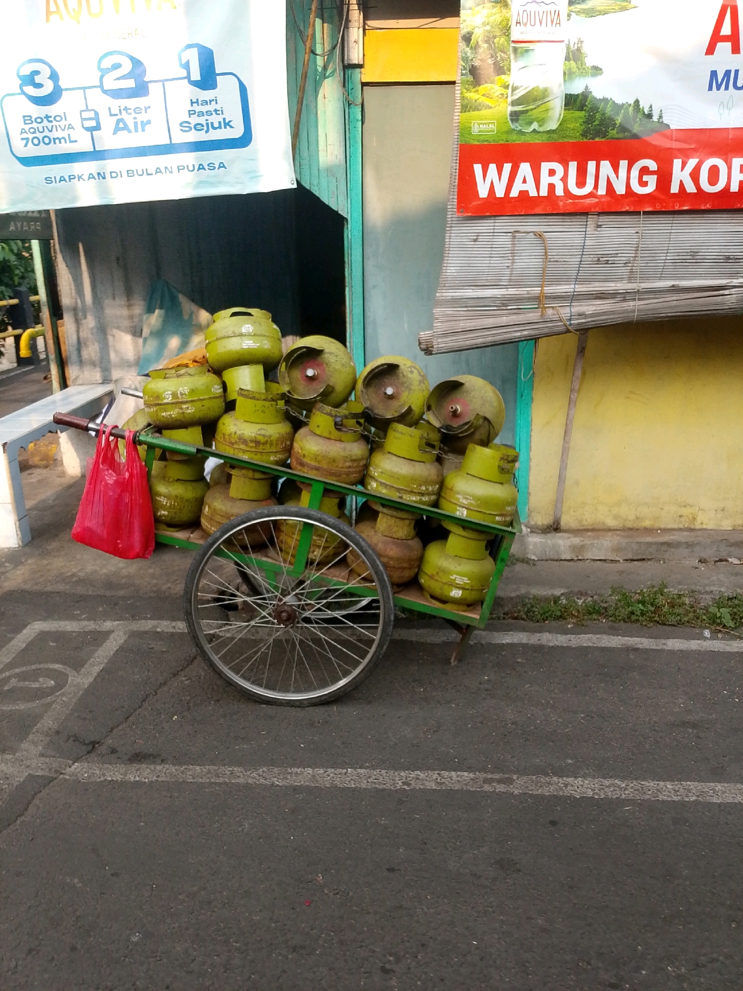 The image captures a street scene in East Jakarta, Indonesia, featuring a cart laden with green LPG cylinders. The cart, equipped with a single spoked wheel, is positioned on an asphalt road marked with white lines. A red plastic bag hangs from the handle of the cart. In the background, a wall painted yellow with a green stripe is visible, along with a bamboo blind and a banner advertising "WARUNG KOP" (Coffee Shop). Another banner in the upper left corner displays text related to "AQUVIVA". The scene appears to be set outdoors, possibly in front of a small business.