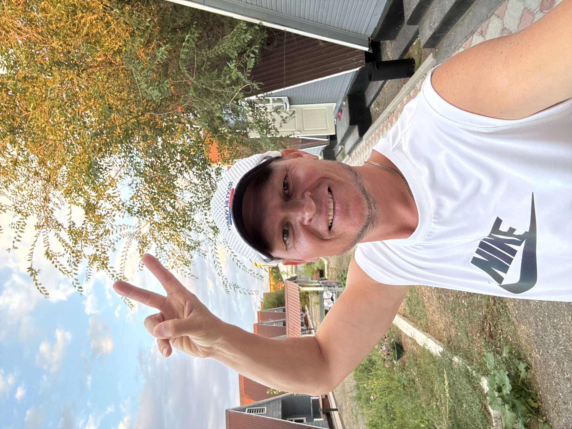 A Caucasian male, appearing to be in his late 30s or early 40s, smiles and holds up two fingers in a peace sign. He is wearing a white sleeveless Nike shirt and a white baseball cap with "TOMMY HILFIGER" written on it. He is standing outside, with a row of small, triangular-shaped houses in the background. The houses have red roofs and grey siding. There is a tree with yellow and green leaves to the right of the man. The sky is visible and appears to be partly cloudy. The ground in front of the houses is paved with small stones. The overall scene suggests a casual, outdoor setting, possibly a resort or a small village. The location is identified as Zharbulak, Kazakhstan.