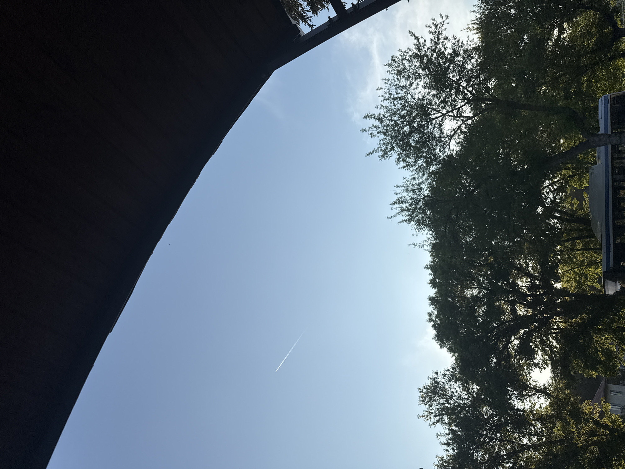 The image depicts a low-angle view of the sky, partially obstructed by a dark, angled structure on the left and trees on the right. The sky is blue with a contrail visible diagonally across the center. A vehicle, possibly a bus, and rooftops are discernible through the trees. The location is Konstantinovo, Bulgaria.