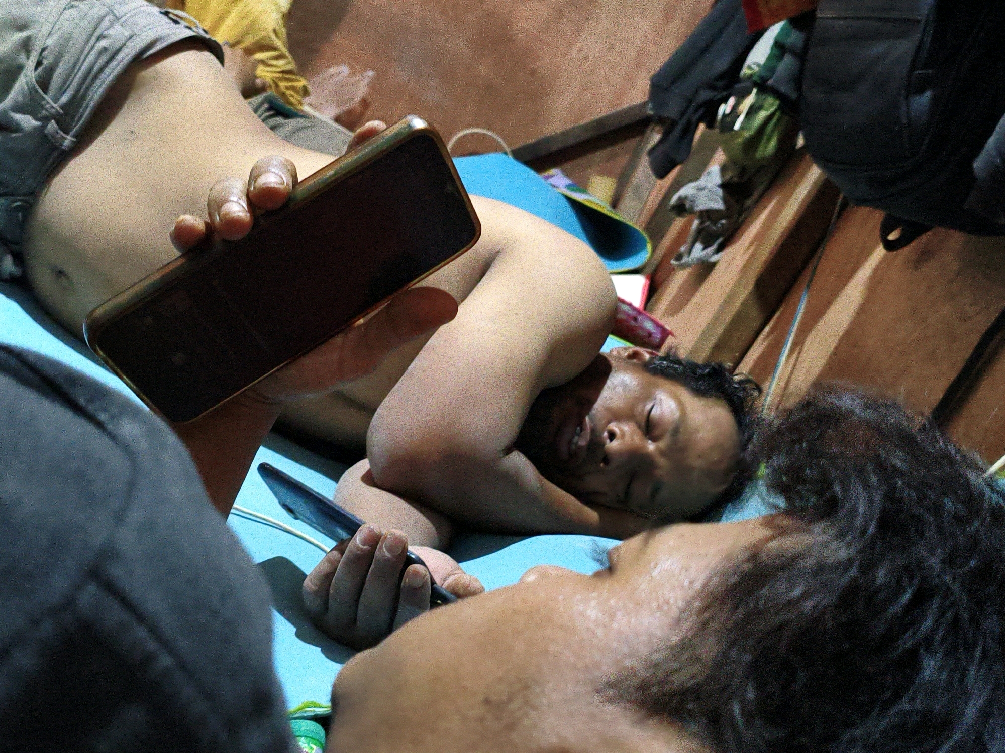 In a close-up, low-angle shot taken in Bekasi, Indonesia, two individuals are seen lying down, possibly on a bed or mat, on a bright blue surface. The person in the foreground, in focus, has dark, curly hair obscuring much of their face, and their head is turned towards the right. Partially visible on the left is a person wearing gray shorts and a yellow shirt. This person is holding a dark-colored smartphone horizontally in their right hand, fingers gripping the sides. The phone's screen is not visible. The person in the background, out of focus, is lying on their back with their eyes closed and head turned to the right. They are partially supported by their arm. The environment suggests an indoor setting with wooden structures and a reddish-brown wall in the background. A black backpack is visible to the right of the frame. The scene is dimly lit, with some ambient light illuminating the subjects.