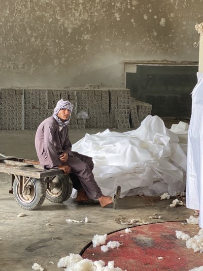 A man sits on a cart in a warehouse filled with stacks of gray blocks and piles of white material. The man wears a headscarf and a long-sleeved, button-down shirt. The floor is concrete with scattered white fluff. The building has bare walls and a loading bay in the background. The setting appears to be a storage area within an industrial building in Kandahar, Afghanistan.