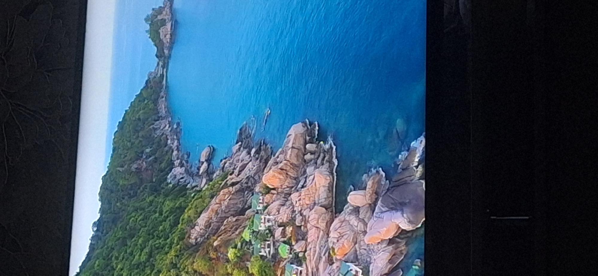 This is an aerial drone shot of a tropical coastline with lush green vegetation covering rocky hillsides. The water is a vibrant blue, with clear turquoise shallows near the shore. Several small, white structures with green roofs are visible nestled amongst the rocks, suggesting a resort or housing development. The rocky shoreline is characterized by large, tan-colored boulders. In the distance, a small, rocky island with some vegetation sits in the sea. The image captures a serene and picturesque coastal landscape. There is no indication of the city of Bila Tserkva, Ukraine, in this image.