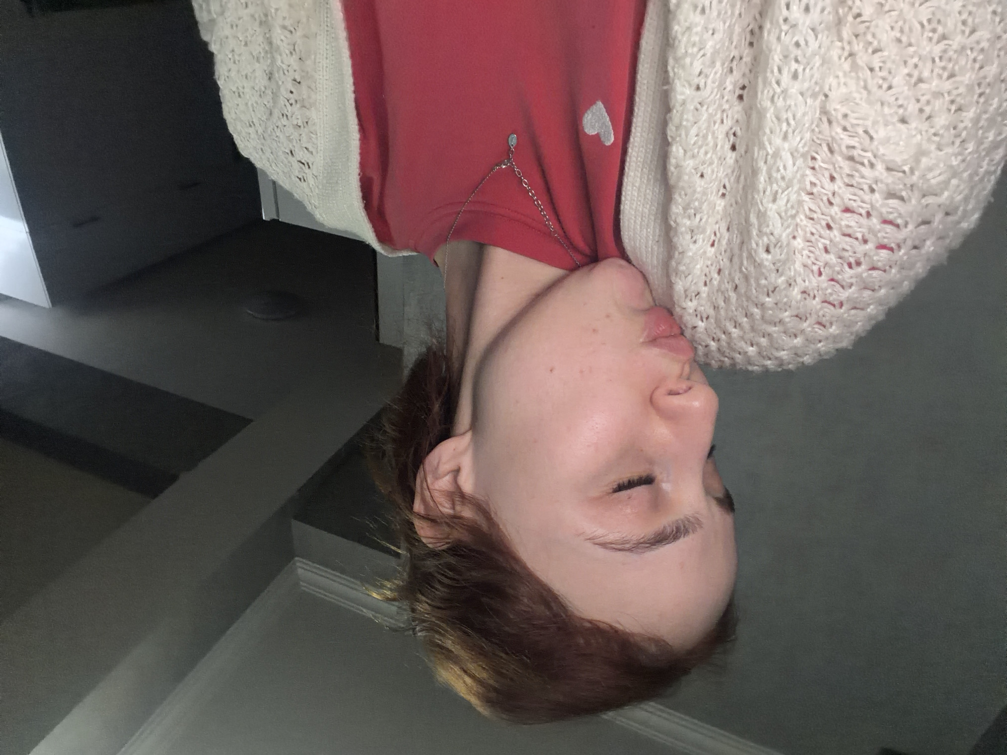 An upside-down, close-up, medium shot captures a young individual from the chest up, set against a dark, indistinct interior background. The person, with fair skin and light brown hair, appears to have their eyes closed or mostly closed, and their lips are slightly parted. They are wearing a red t-shirt that features a small, light grey heart emblem on the left chest area. Over the t-shirt, a light-colored, possibly cream or off-white, knitted cardigan with a distinct textured pattern is visible. A delicate silver-colored chain necklace, adorned with a small, tear-drop shaped pendant, rests on the red t-shirt. The background consists of dark, solid surfaces, likely interior walls or furniture, with subtle lighting suggesting an indoor setting. The individual is posing for the photograph, conveying a calm or contemplative emotion due to their closed eyes. There is no visible text in the image.