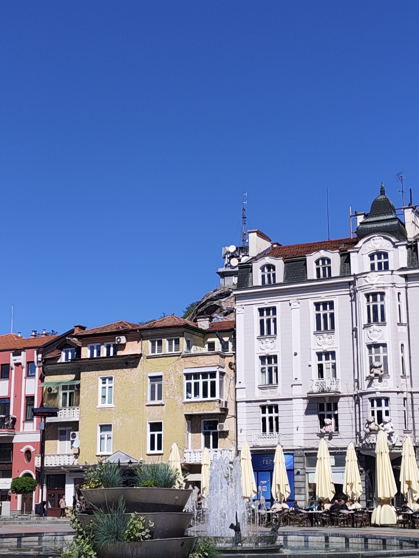 A street scene in Plovdiv, Bulgaria, features colorful buildings along a square with a fountain in the foreground. Several buildings are visible, ranging from pink to yellow to white, each with multiple windows and balconies. A fountain with several levels of tiered planters filled with greenery sprays water in front of the buildings. Cream-colored umbrellas shade tables with seated people, suggesting a café or restaurant area. A hill or rock formation is visible behind the buildings, topped with a structure that appears to be a tower or monument. The sky is clear and blue.