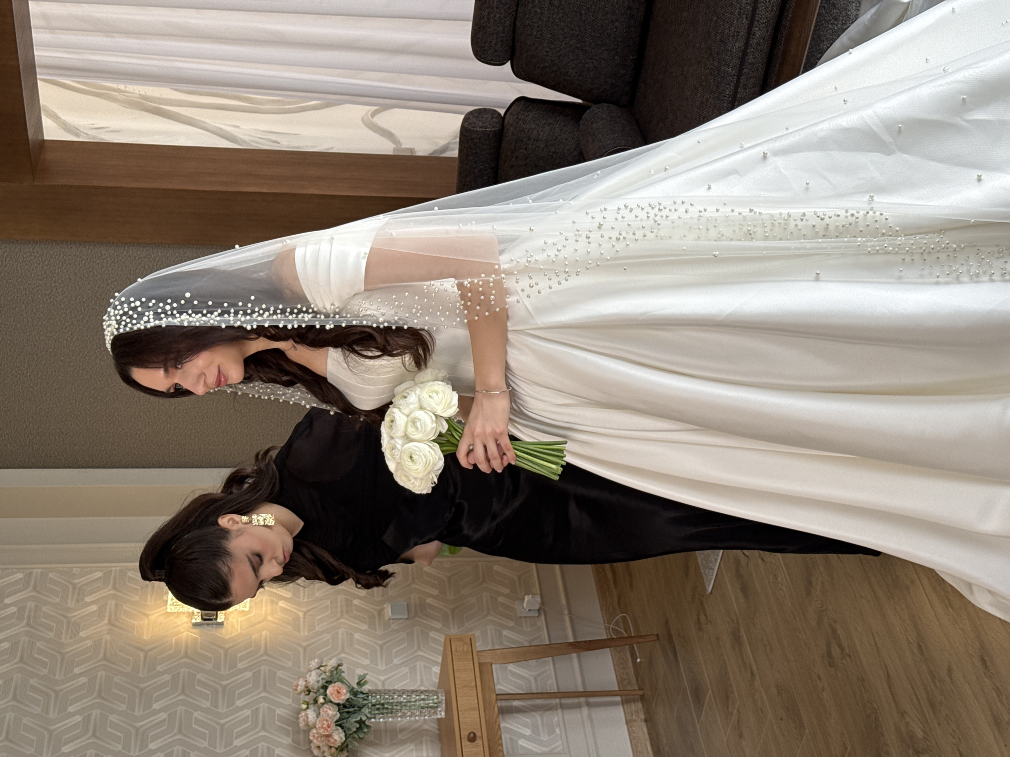 The image displays two women standing indoors, likely in Makhachkala, Russia, during a wedding event. The woman on the right is a bride, wearing an elegant white satin wedding gown and a long, sheer veil meticulously adorned with scattered pearls. She holds a lush bouquet of white ranunculus flowers in her hands, and a delicate bracelet is visible on her left wrist. She has dark hair and a pensive expression, looking downwards.
Beside her, on the left, stands another woman in a formal long black dress, also with dark hair styled in soft waves. She wears distinctive gold-toned, geometric earrings. Her gaze is similarly directed downwards, suggesting a shared moment of focus or contemplation.
The setting is an interior space, featuring light-colored patterned wallpaper with a subtle geometric design and a warm wooden floor. A wall sconce provides lighting, and dark upholstered armchairs are partially visible in the background. In the foreground, a wooden stand or table holds a vase with light pink and white flowers. The atmosphere is calm and intimate, capturing a quiet moment between the two women. There is no visible text in the image.