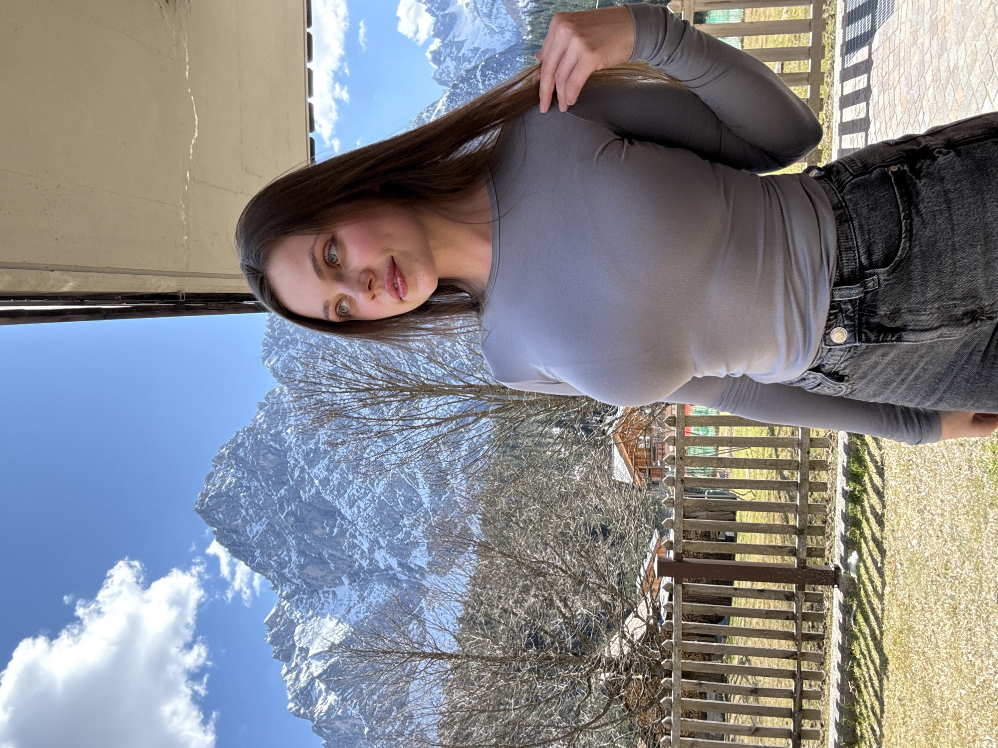A woman in a gray long-sleeved shirt and dark jeans stands on a balcony. She is running her fingers through her long brown hair and looking at the camera. In the background is a mountain range with snow on the peaks and a blue sky with clouds. Below the mountains is a wooded area with bare trees. In the foreground is a wooden railing with shadows cast on the ground. The location is in Sappada / Plodn / Sapade, Italy.
