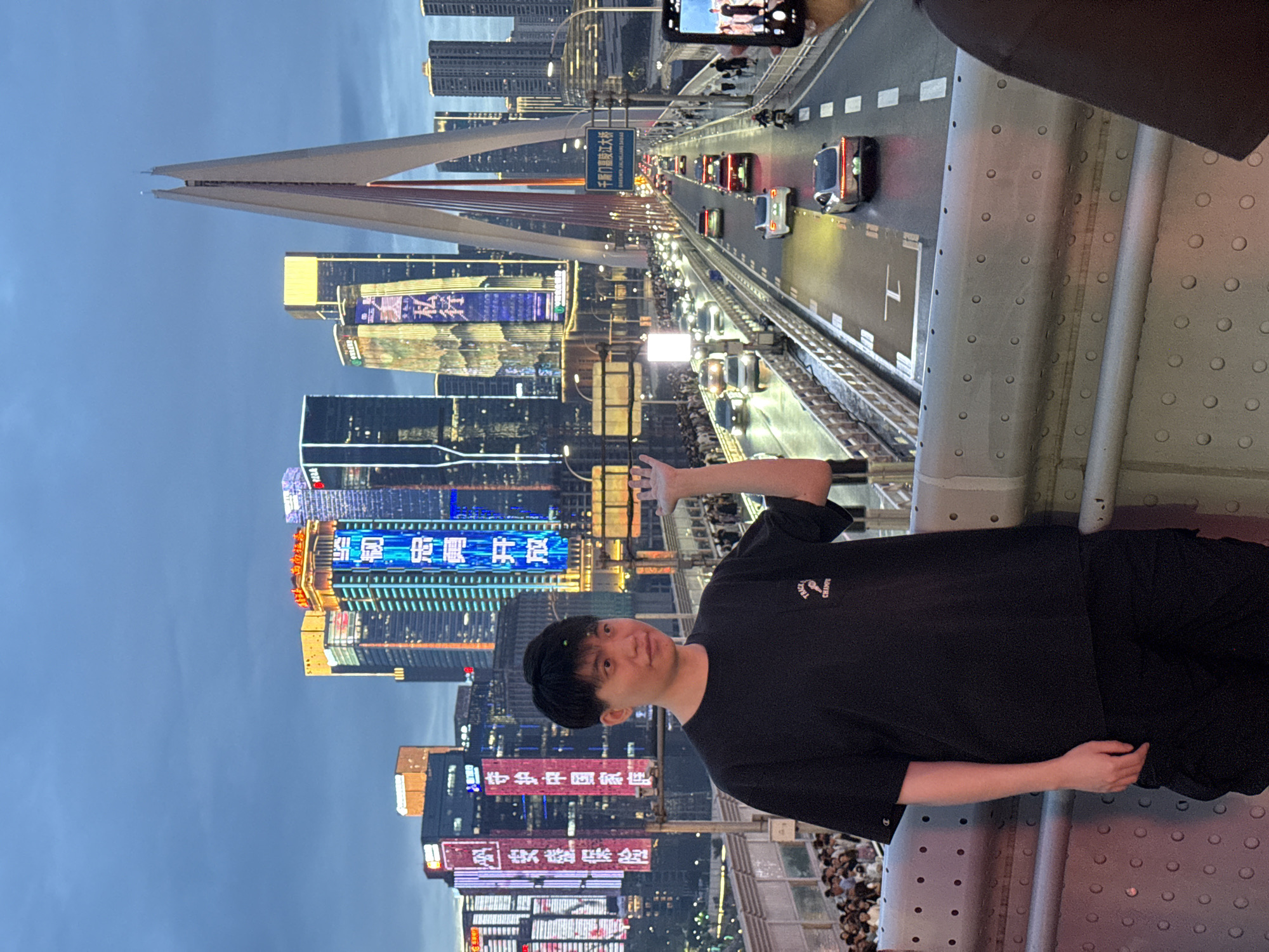 A young man with short black hair stands on a bridge, facing the camera, with a cityscape in the background. He is wearing a black t-shirt with a logo on the left chest and black cargo pants. His right hand is raised and extended outward, palm facing the viewer. Behind him, skyscrapers with illuminated signage light up the dusk sky. The bridge appears to be a busy thoroughfare, with traffic visible on the roadway below. Cars, motorcycles, and pedestrians are present. In the foreground, a metal railing with circular indentations runs along the edge of the bridge. On the right side of the frame, a hand is visible holding a smartphone, with a portion of another person's dark shirt in the lower right corner. The scene is set in the Yuzhong District of Chongqing, China, likely on the Hongyadong Bridge, given the prominent architecture.