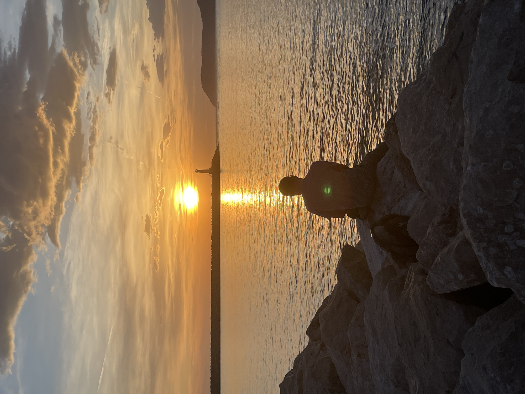 A person is sitting on large rocks, facing away from the camera, toward a sunlit body of water at sunset. The sun is setting behind a distant structure that appears to be a lighthouse or tower. The sky has orange and gold hues with clouds visible to the left side of the frame. The water reflects the sunlight creating a bright path to the distant structure. The rocks are grey and rough in texture. The location is in the city of Bar, Montenegro.