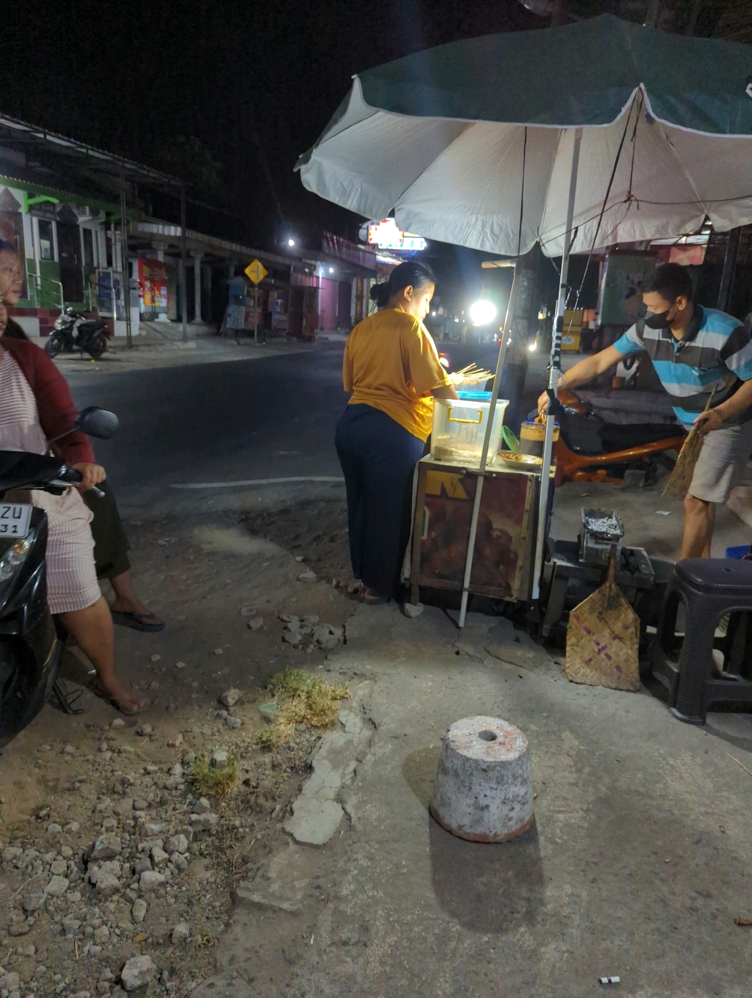 The image depicts a night scene on a street in Kacangan, Indonesia. A woman in a yellow shirt and dark pants stands behind a food cart, attending to skewers of food. A man wearing a blue and black striped shirt and a mask is on the right side of the cart, holding a broom and seemingly engaged in cleaning or organizing. A large, partially open umbrella, white on the inside and green on the outside, is set up over the food cart. In the foreground, to the left, a motorcycle is partially visible, with a woman in a pink and white striped dress and a maroon jacket seated on it. The street appears to be paved, with a rough, uneven area of gravel and dirt to the left of the cart. In the background, buildings line the street, illuminated by artificial lights. The overall impression is that of a typical street food vendor operation at night in a semi-urban Indonesian setting.