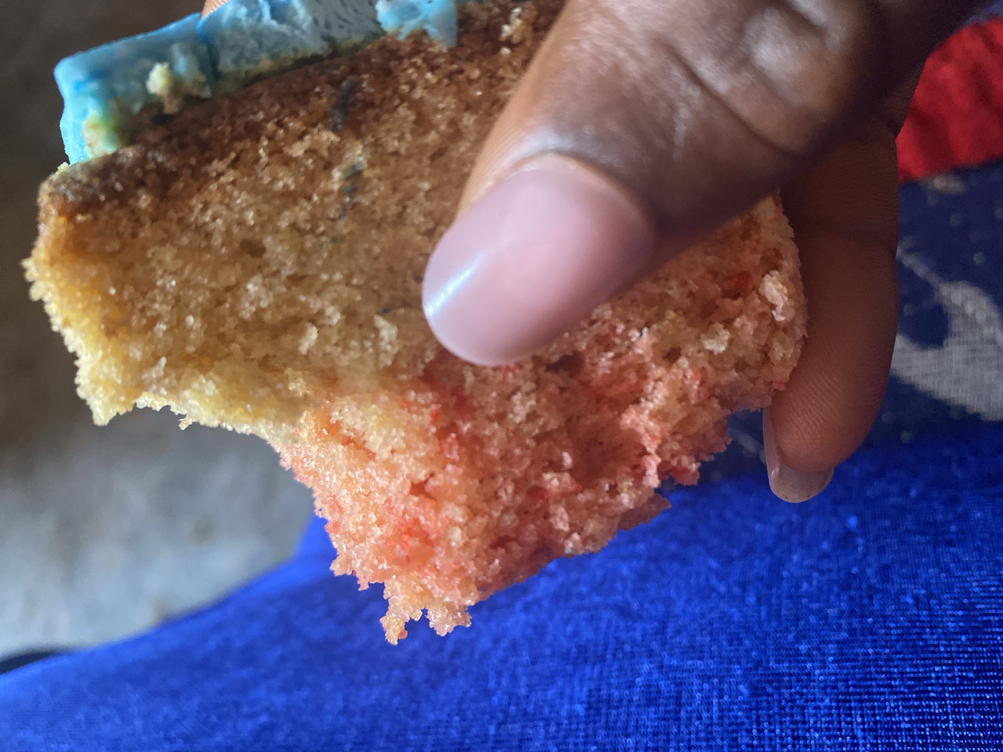 The image shows a close-up of a person's hand holding a piece of cake with blue frosting and pink cake. The person's hand is dark-skinned, and their fingernail is visible and appears to be manicured with a pinkish hue. The cake appears to be a slice from a larger cake, with a crumbly texture. The background is blurred, but it appears to be a blue fabric. The image does not provide any information about the location in the city of Wumat, Nigeria.