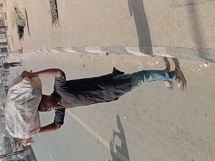 The image depicts a daytime scene on a street in Sharada, Nigeria. A young adult male, positioned left of center, is standing on the paved roadside next to a concrete curb. He is wearing a dark, long-sleeved tunic, faded blue jeans, and light-colored slide sandals. He balances a large, off-white, irregularly shaped sack on his head, supporting it with both arms raised. His gaze is directed forward.
The foreground and midground consist of a worn asphalt or concrete road surface with visible debris. A concrete curb separates the road from a dirt shoulder on the right, which contains an open drainage ditch or culvert section and additional scattered debris. In the background, a multi-lane road extends into the distance, where several individuals, "keke napep" (tuk-tuks), and other vehicles are present. Strong shadows cast by the curb and unseen objects indicate bright daylight conditions.