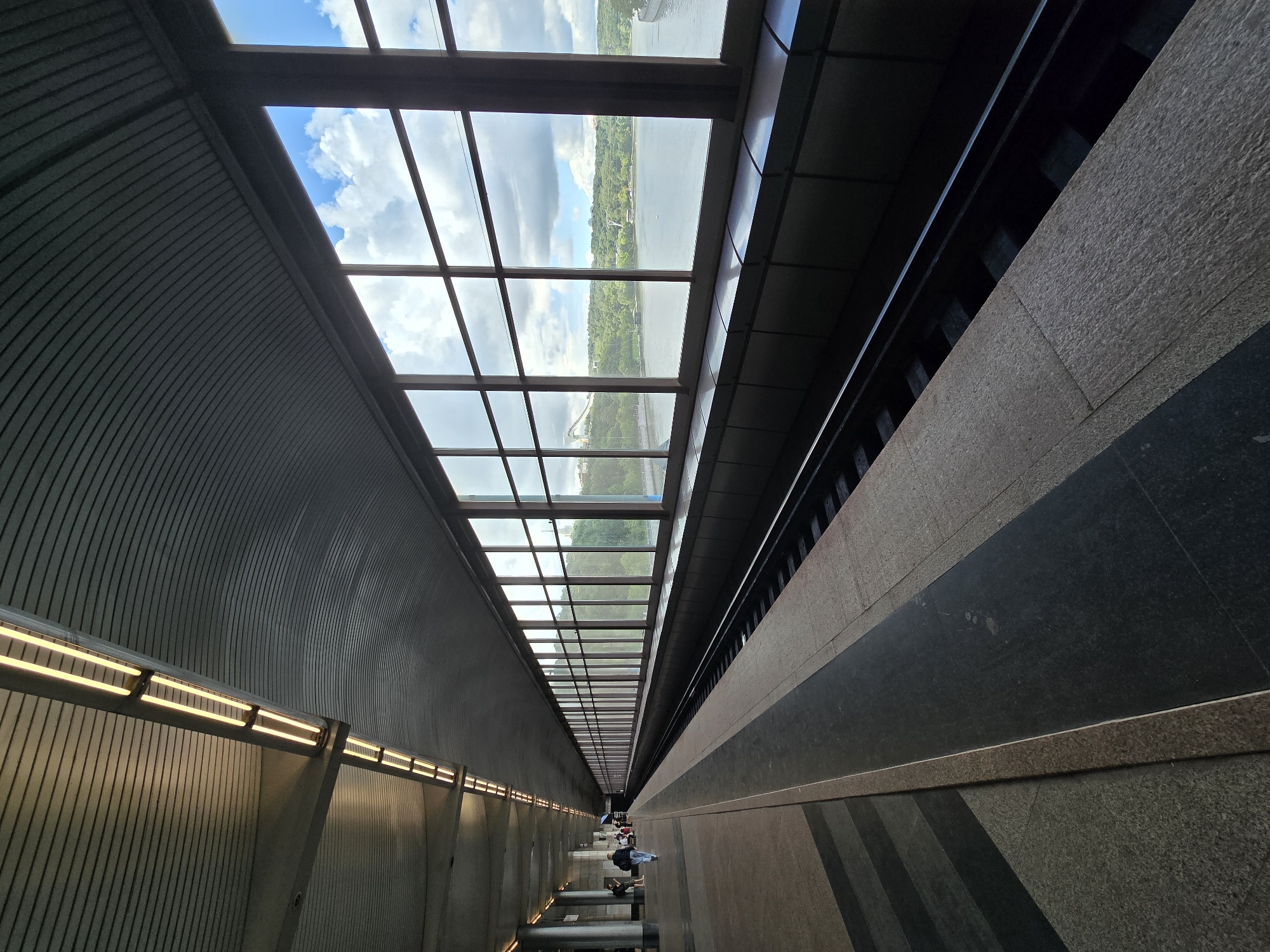 An outdoor, elevated metro station platform with a long row of windows is pictured. The platform is made of granite tiles with a dark grey border. The windows overlook a wide river with green hills and a bright blue sky with fluffy white clouds. The station is covered by a curved, metal ceiling with rows of fluorescent lights. A set of train tracks runs along the platform. In the distance, on the left side of the image, a few people are walking and standing on the platform.