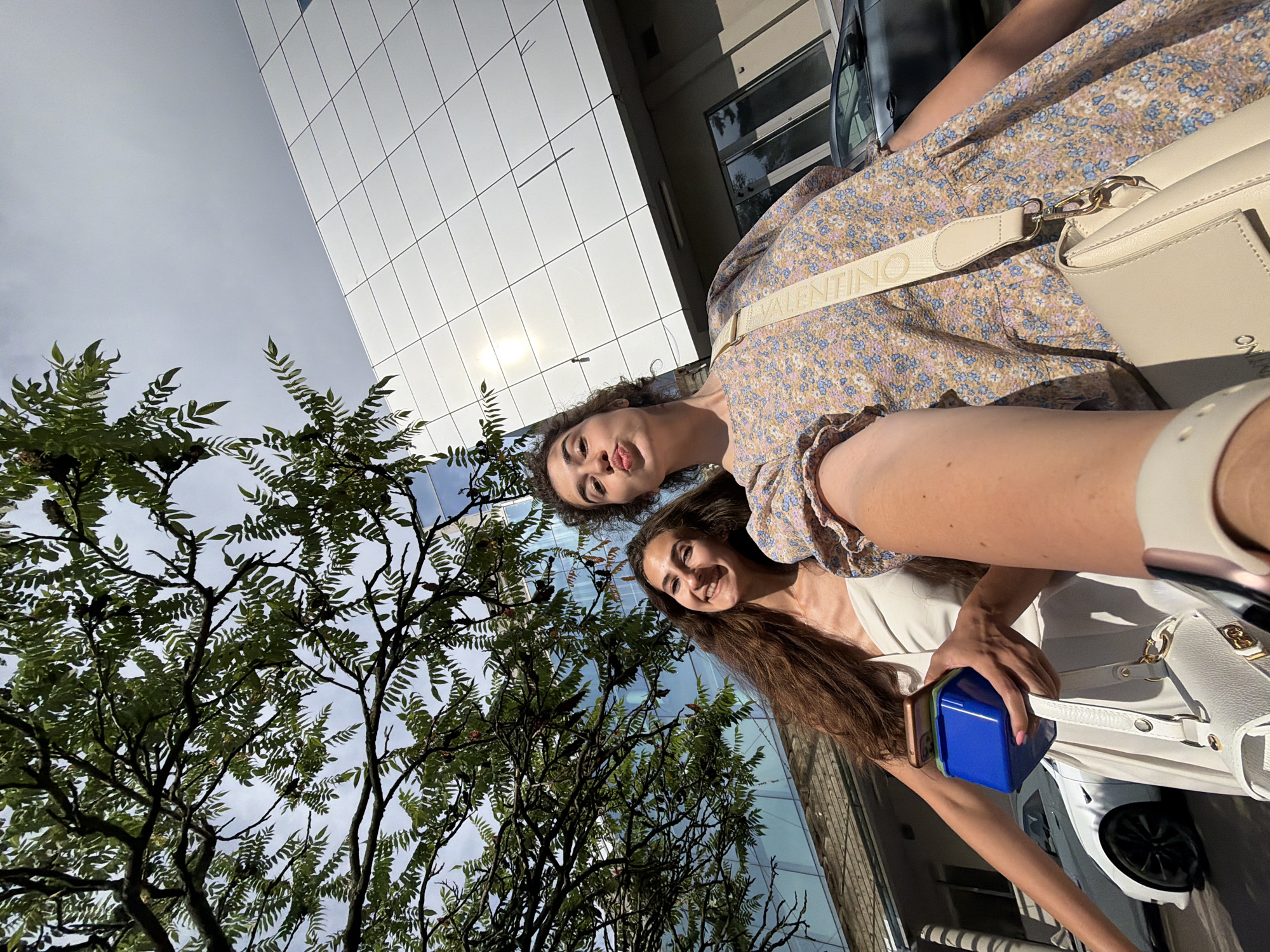 This media file is a selfie taken outdoors, likely in an urban environment. Two young women are the primary subjects, positioned in the foreground and mid-ground of the image. The woman in the foreground, on the right, has long, brown hair and is smiling. She is wearing a light-colored dress and a cream-colored handbag with a prominent strap that has the word "VALENTINO" printed on it in light text. The second woman, in the mid-ground on the left, has curly dark hair and is making a playful face, sticking out her tongue. She is wearing a floral print dress.
The setting includes modern architecture with a building facade composed of white, grid-like panels reflecting the sky. To the left of the frame, there are lush green trees with dense foliage and branches. The sky is visible, appearing overcast with hints of sunlight breaking through. The time of day appears to be daytime, possibly late afternoon, given the angle of the sunlight and shadows. The overall mood seems cheerful and casual. One of the women is holding a small blue rectangular object and two mobile phones.
Visible text: "VALENTINO".
Location context: Sofia, Bulgaria.