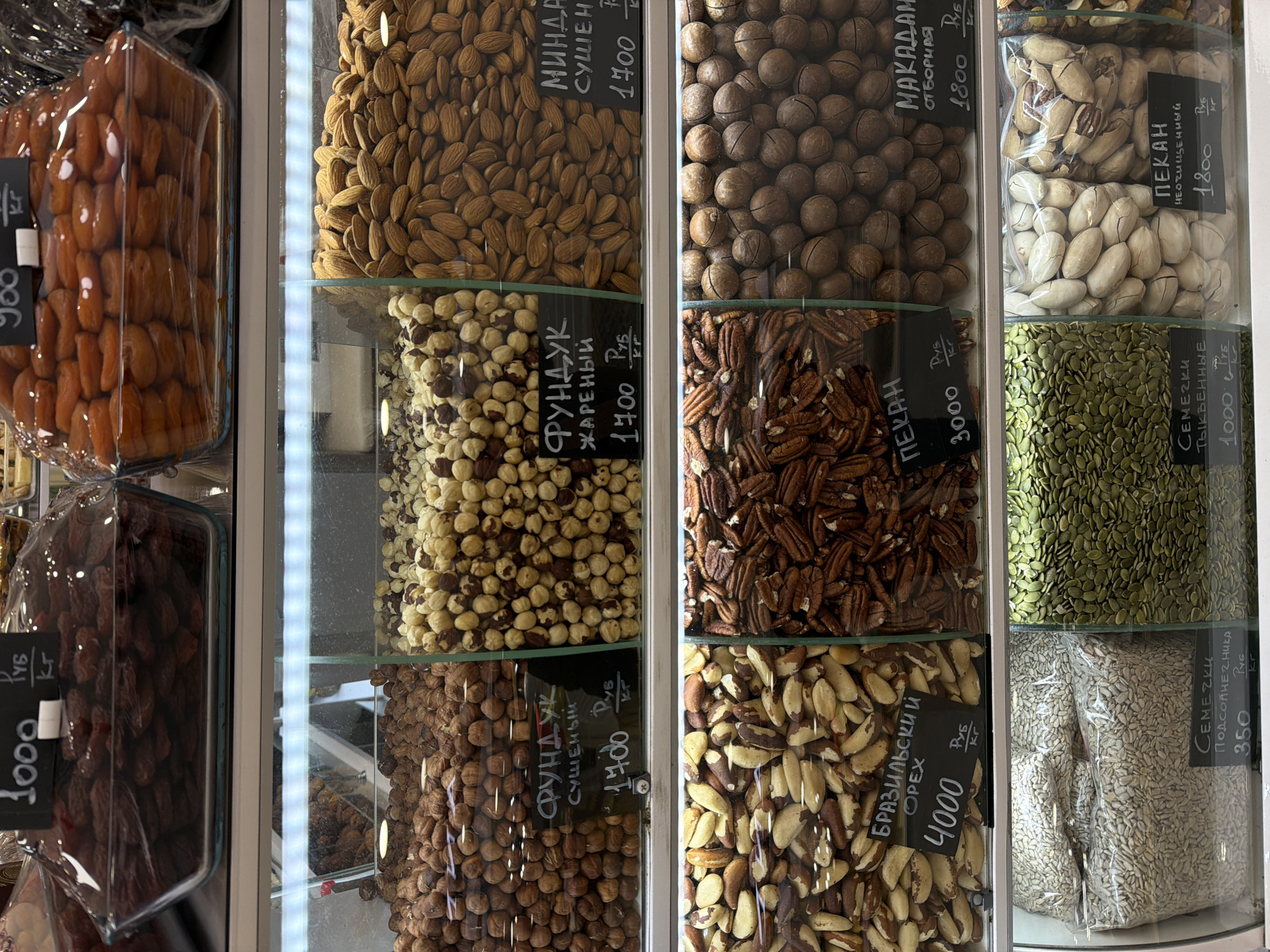 This image shows a close-up view of a display case filled with various types of nuts, seeds, and dried fruits. The display seems to be within a store in Moscow, Russia, suggested by the Russian text on the labels.
The nuts and seeds include almonds, hazelnuts (both raw and roasted), macadamia nuts, pecans (both shelled and unshelled), Brazil nuts, pumpkin seeds, and sunflower seeds. There are also dried apricots and prunes. Each section of the display has a label indicating the type of product and its price per kilogram in Russian rubles. The prices range from 350 to 4000 rubles.
The display case is brightly lit, which helps highlight the textures and colors of the different products. The items are arranged in separate sections with clear containers. The overall impression is a visually appealing assortment of snack and food items.