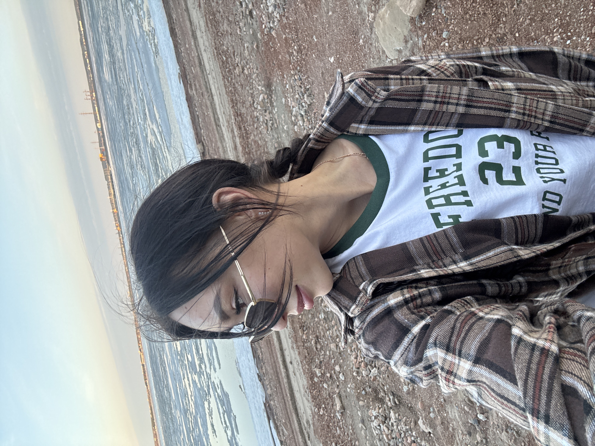 A young woman stands outdoors, likely near a body of water. She is wearing a white t-shirt with green text and numbers, a brown plaid shirt, and gold-framed sunglasses. Her dark hair is pulled back. The background shows a shoreline with ice or snow on the water and what appears to be a cityscape in the distance. Given the mention of Noviy, Russia, it may be the coast of a river or lake during a cold season. The ground appears to be gravel or dirt.