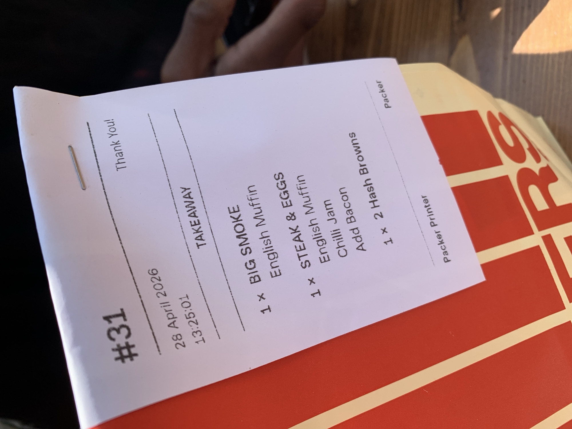A close-up, diagonal shot captures a white paper takeaway receipt resting on a red, branded box or bag, with blurred fingers visible in the background. The scene is brightly lit, likely indicating daytime.
The receipt details a specific food order. Prominently displayed is "#31" at the top left, followed by the date "28 April 2026" and time "13:25:01". The order is designated as "TAKEAWAY". The items listed are:
* "1 x BIG SMOKE" with an accompanying "English Muffin".
* "1 x STEAK & EGGS", further customized with an "English Muffin", "Chilli Jam", and "Add Bacon".
* "1 x 2 Hash Browns".
Additional text on the receipt includes "Thank You!" at the top, and "Packer Printer" and "Packer" along the right side, suggesting an internal processing record. A small metal staple is visible on the receipt.
The red object beneath the receipt features a distinctive white grid pattern and the partial letters "RS", hinting at branded packaging from a food establishment. The overall impression is of a prepared food order ready for collection or delivery, consistent with a quick-service restaurant or cafe in a setting like London, UK.