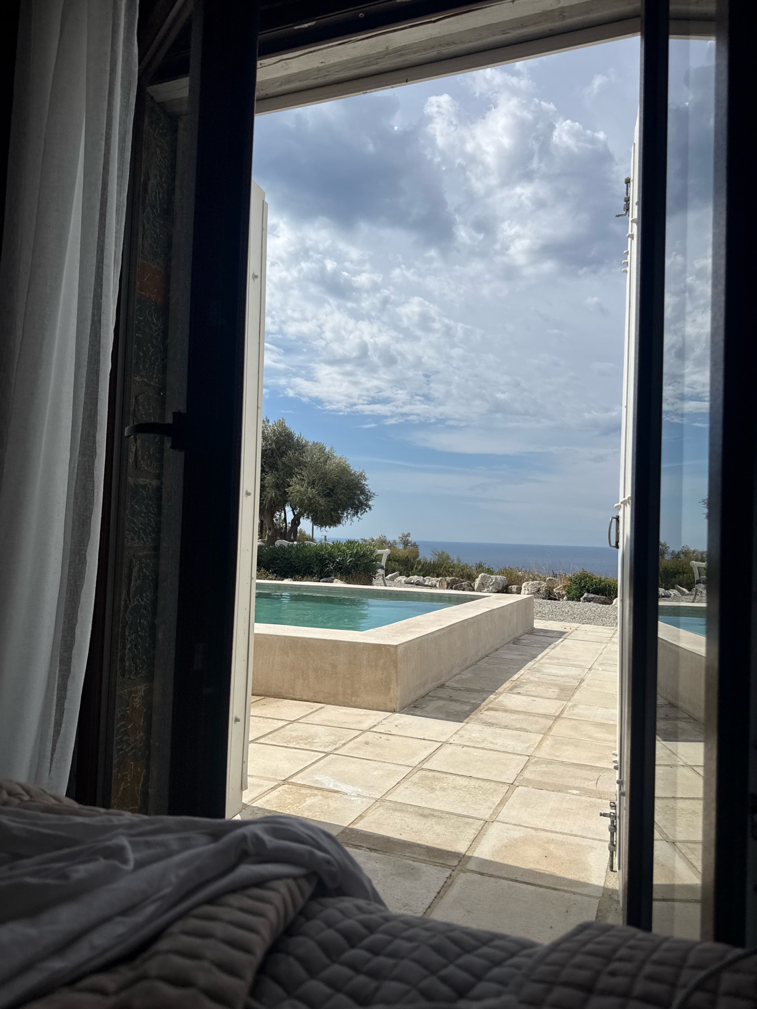 This is a medium shot, taken from inside a room, looking out through an open doorway towards a swimming pool and the sea. The room has a white curtain on the left side and a quilted blanket in the foreground. The doorway has dark frames.
Outside, a light blue swimming pool with a beige border is visible. The pool area is paved with large square tiles. Beyond the pool, there is a rocky landscape with sparse vegetation and a large tree. In the distance, the blue sea stretches to the horizon under a cloudy sky with white and grey clouds.
The scene appears to be set in Mandres, Greece.