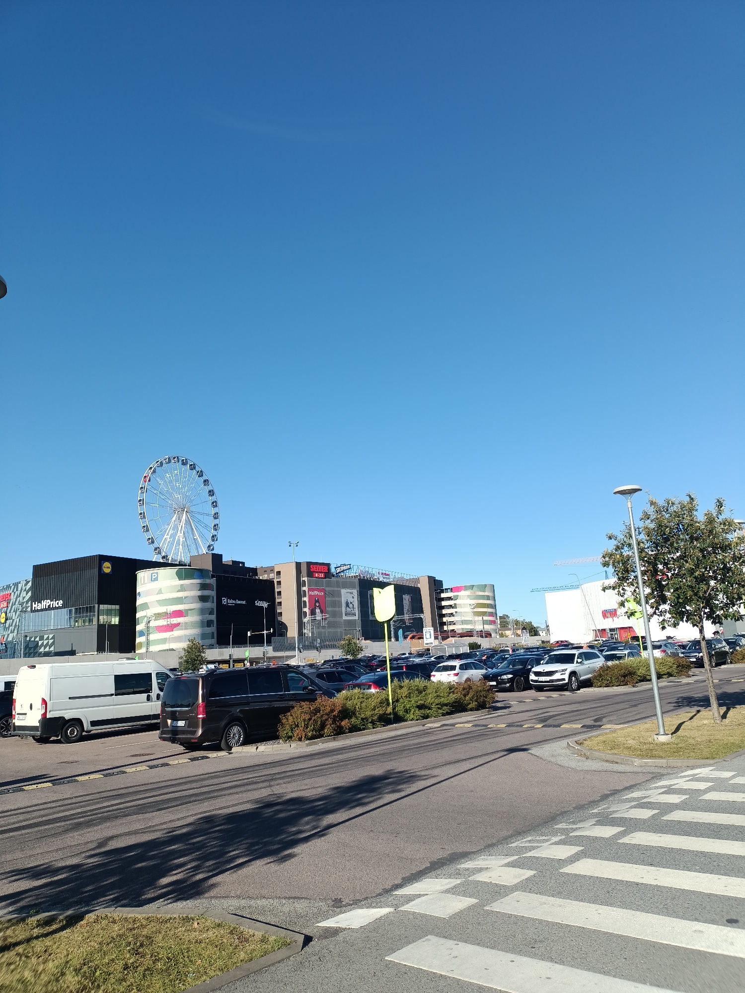 The image shows a view of the Ulemiste City shopping complex in Tallinn, Estonia, under a clear blue sky. A large Ferris wheel is visible behind the buildings. In the foreground, a paved road leads to a parking lot filled with various cars, including a white van and a dark minivan. Some of the vehicles are parked in designated spaces, while others appear to be in traffic lanes. Bushes and trees line the edges of the parking lot and the road. Shadows of trees are cast across the asphalt. A pedestrian crossing with white stripes is prominent in the lower right corner of the image. The buildings in the background display various signs, including "HalfPrice," "Lidl," and "SEB." The overall scene depicts a commercial area on a sunny day.