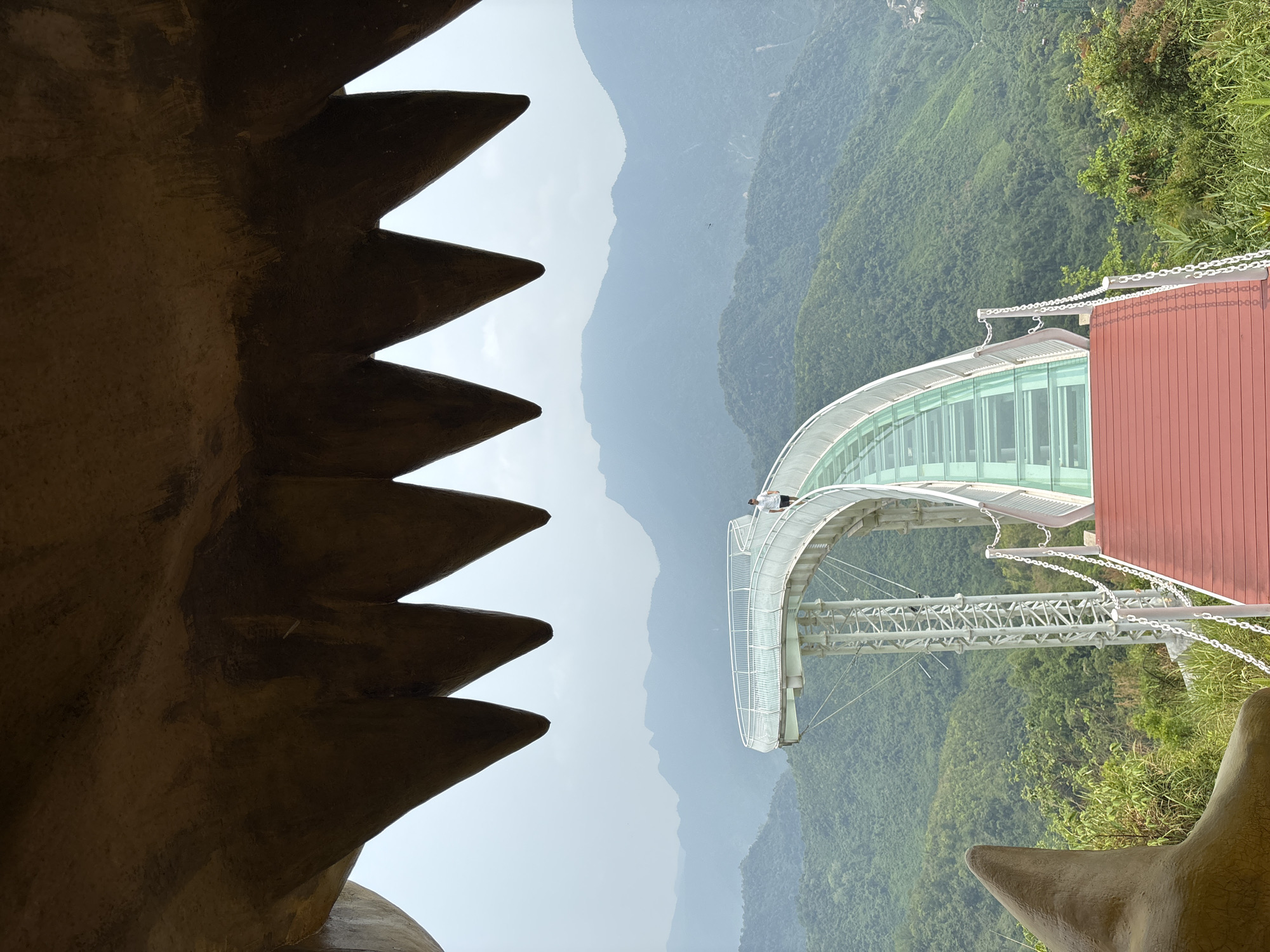 The image shows a view from inside a structure with jagged edges, possibly representing teeth, overlooking a mountainous landscape in Đà Nẵng, Vietnam. A curved glass-enclosed walkway is visible, extending into the lush green mountains. The sky is overcast. A person is standing on the walkway. The walkway appears to be part of a tourist attraction.