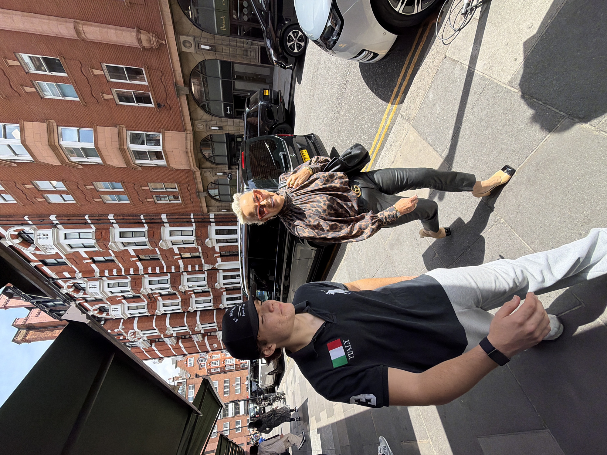 In London, United Kingdom, two individuals are walking on a sidewalk adjacent to a street lined with tall, red brick buildings featuring white decorative trim and numerous windows. A man in the foreground, wearing a black polo shirt with an Italian flag patch and the word "ITALY" below it, along with light grey sweatpants and a black baseball cap, is looking to his right. He has a black fitness tracker on his left wrist. To his right and slightly behind him, a woman with blonde hair styled in a blonde bun, wearing large, red-rimmed sunglasses, a leopard-print blouse with a high neckline, and black leather pants, is walking and looking towards the camera. She has a black handbag slung over her left shoulder and is wearing pointed-toe, two-toned heeled shoes. Behind the individuals, several vehicles are parked along the street, including a black van and two SUVs. The buildings appear to house storefronts, with one visible sign reading "Harrods." The sun casts shadows on the sidewalk, indicating it is daytime.
