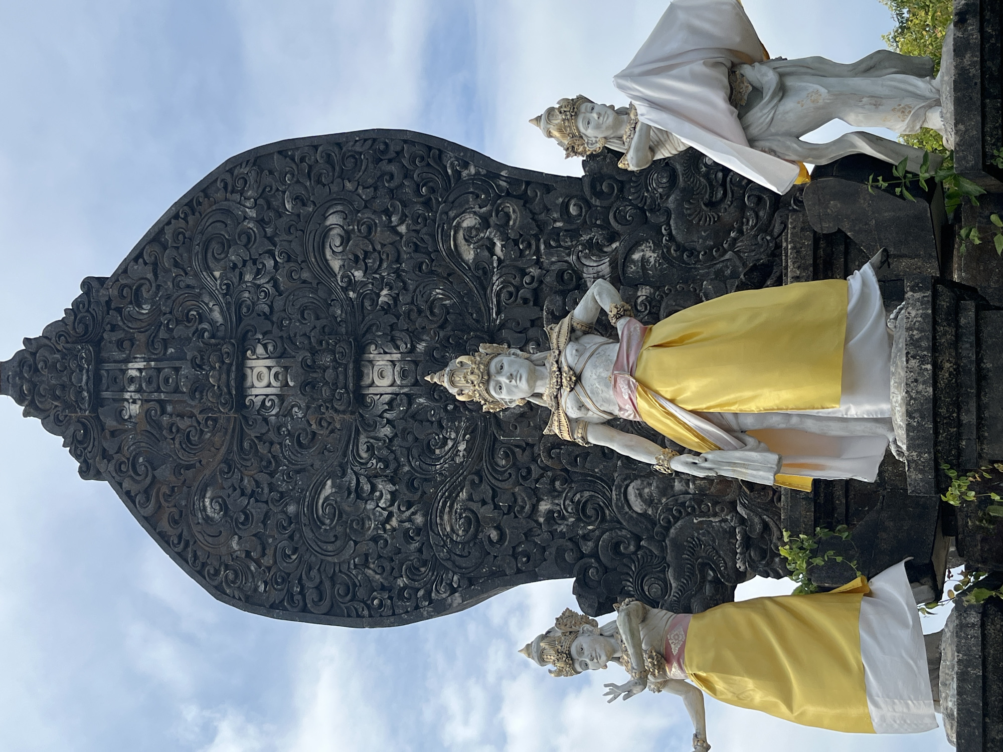 In Kuta Selatan, Indonesia, a trio of statues stands before a large, ornate, dark-colored stone carving. The central statue, a female figure, is adorned with a golden crown and a yellow sash over a white garment. To her left, another female statue wears a similar crown and a yellow sash with a white underskirt. To the right, a third female statue, also crowned, is partially obscured by white fabric draped over her arm and body. The statues are positioned on stone pedestals, with greenery growing around the base. The background features a cloudy blue sky.