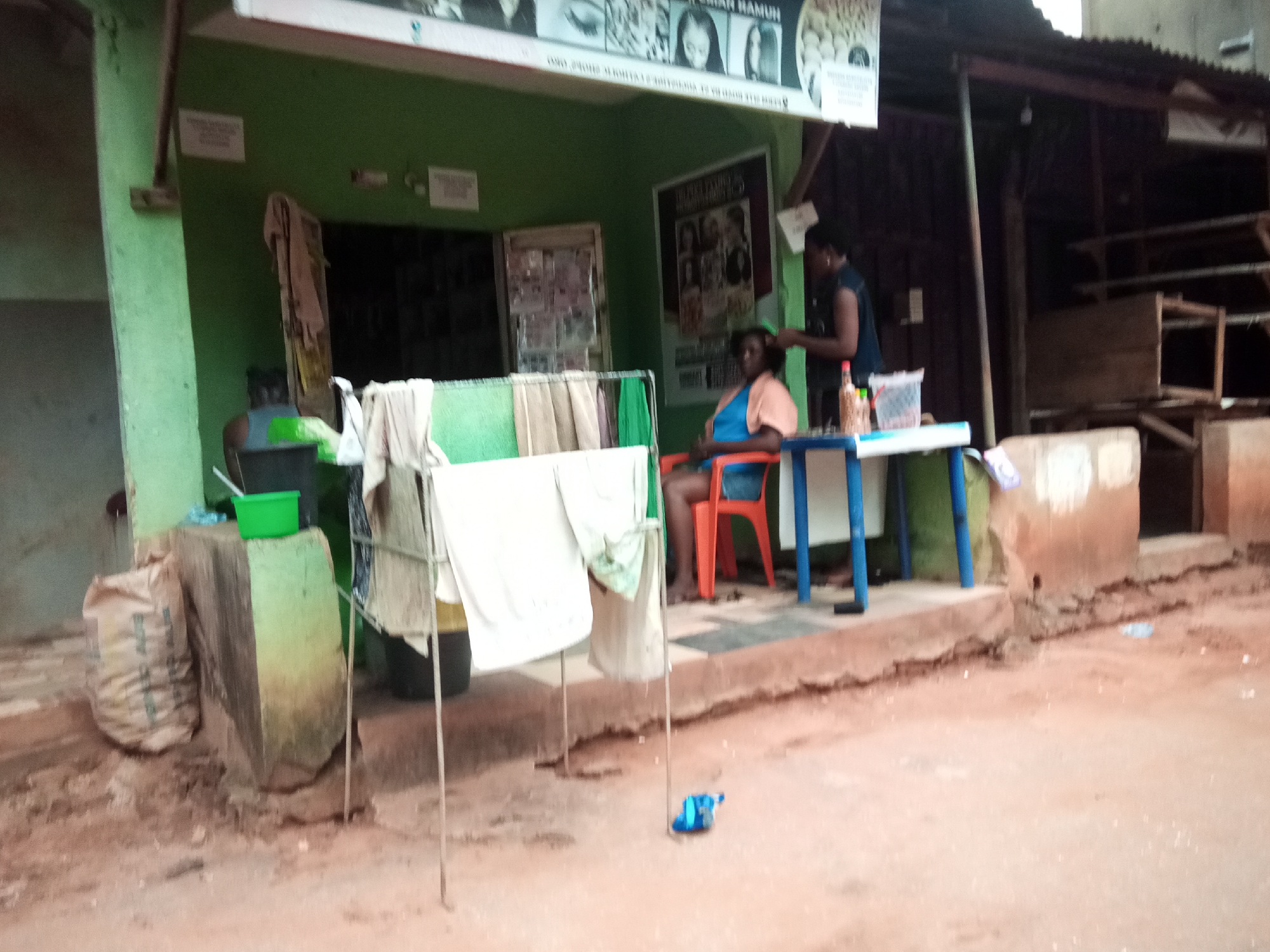 In Oko Town, Nigeria, a woman is getting her hair styled by another woman inside a small, open-air shop. The shop has a green facade with various posters and signage displayed on the wall. The woman receiving the hairstyle is seated in an orange chair, wearing a light orange top and a blue dress. The hairstylist, wearing a dark sleeveless top, is actively working on her hair with a comb. In front of the shop, there is a table with a blue frame and a white top, holding some items like bottles and containers. To the left of the shop, a large sack of what appears to be goods is placed next to a concrete structure. Clothes are hanging on a rack in front of the shop. The ground is covered in dirt and sand, with some scattered debris. The overall scene depicts a typical street-side hair salon operation in a bustling Nigerian town.