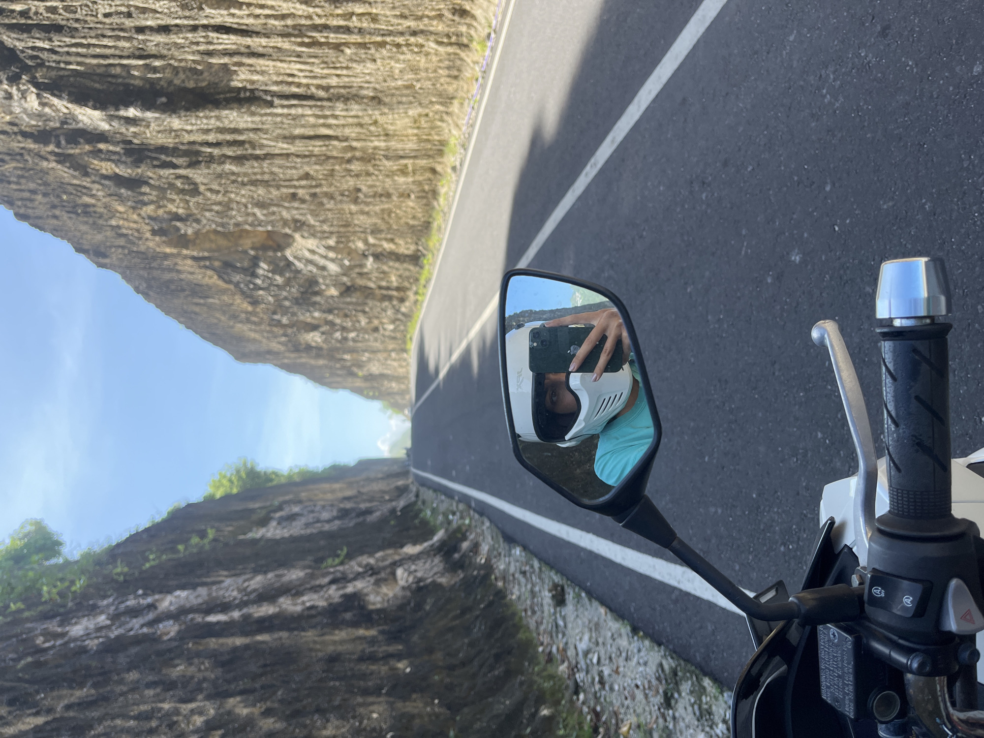 The image shows a motorcycle side mirror reflecting a person wearing a white helmet and a teal shirt taking a selfie. The surrounding environment consists of a road flanked by tall, layered rock formations, suggesting a canyon or cliffside road. The asphalt road has a white line marking the lane. The motorcycle's handlebars and controls are visible in the foreground. The location is likely Kuta Selatan, Indonesia, based on the distinctive geological features often associated with that region.