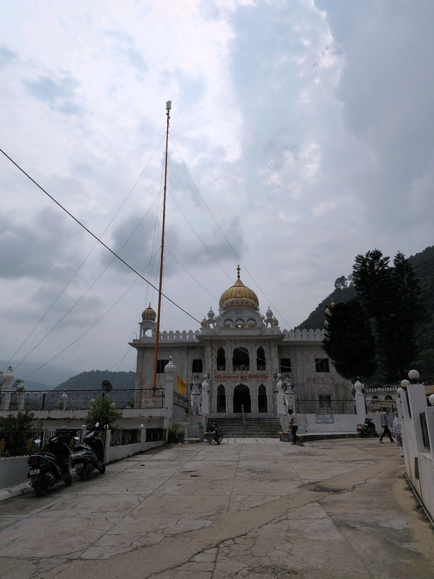 A wide shot captures a Sikh Gurdwara, likely in Mandi, India, under an overcast sky with dramatic clouds. The building features white marble, a golden dome, and intricate architectural details with arched windows and decorative white crenellations. In front of the Gurdwara, a tall, slender flagpole, known as a Nishan Sahib, stands prominently, with a modern horn or speaker attached near its top.
The foreground is a paved courtyard with visible cracks and wear, leading towards the Gurdwara's steps. Several parked scooters and motorcycles are visible on the left side of the courtyard, suggesting a place of public access. In the middle ground, a few individuals are seen walking and interacting within the courtyard area. One person is standing near the Gurdwara entrance, while others are further away, moving towards the right of the frame.
The background reveals lush green hills and trees, indicative of a mountainous or hilly region. The overall impression is a serene, yet active, spiritual site nestled within a natural landscape. The lighting is diffused due to the cloudy conditions, creating soft shadows.