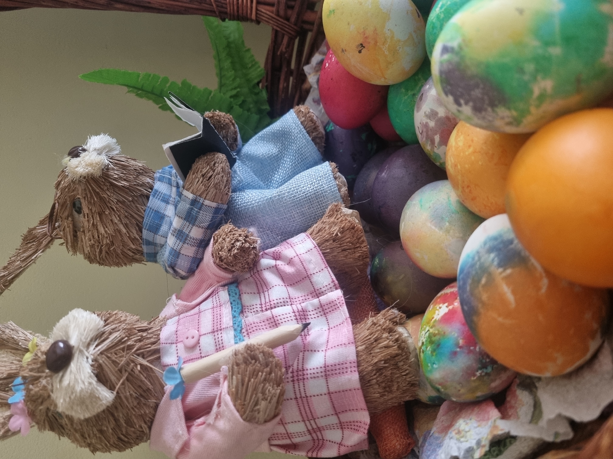 The image shows a wicker basket filled with decorated Easter eggs and two straw bunny figurines dressed in fabric clothing. The eggs are painted in various colors, including yellow, red, green, purple, and orange, with some having a mottled or tie-dye appearance. One bunny is wearing a blue plaid shirt and holding a small book, while the other wears a pink plaid dress and holds a pencil. Green ferns are visible behind the bunnies. The setting appears to be indoors with a neutral-colored wall in the background. The location and city are unspecified.