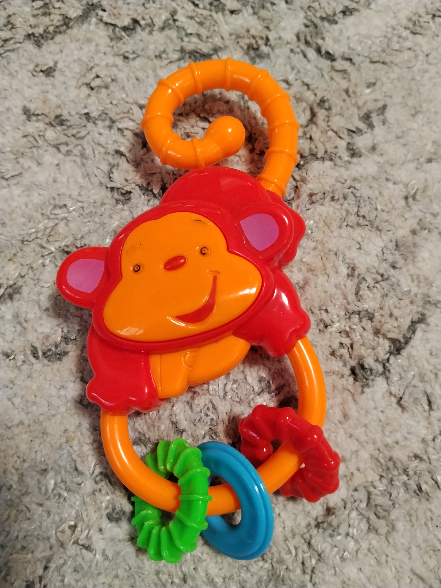A colorful plastic toy, designed as a monkey face with a coiled tail, lies on a grey, textured carpet. The monkey's face is orange with pink inner ears, and its body and tail are red and orange respectively. Attached to the bottom of the toy are several rings: one green, one blue, and another red, each with a different textured surface. The scene is illuminated from above, casting subtle shadows. The specific location of Буренка, Russia is not visually discernible from the provided image.