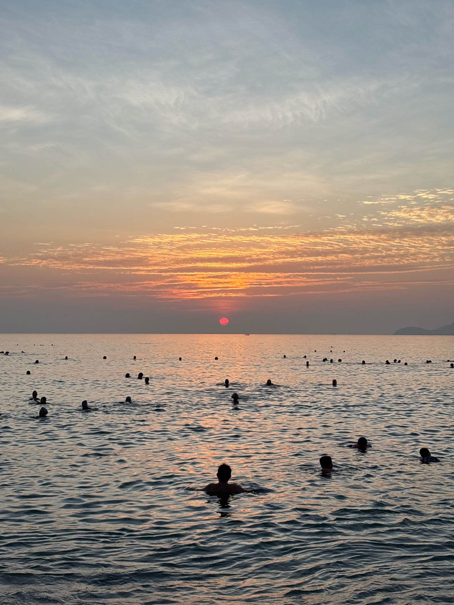 The image captures a serene sunset over the ocean in Nha Trang, Vietnam. A group of people are swimming and wading in the calm sea, their silhouettes visible against the warm hues of the setting sun. The sky is painted with soft orange and pink clouds, creating a picturesque backdrop. The water is gently rippling, reflecting the colors of the sky. In the distance, a faint outline of hills can be seen. The scene suggests a peaceful evening at a beach in Nha Trang.