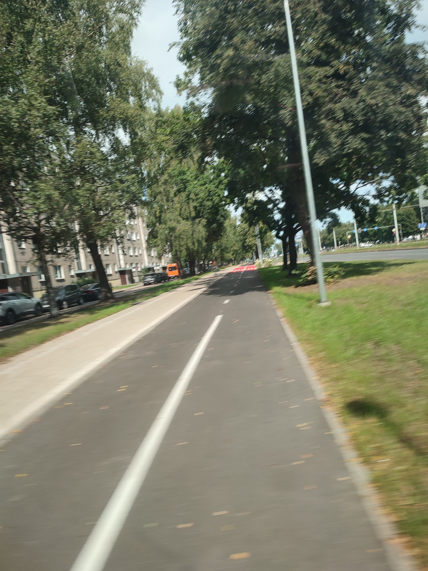 The image depicts a paved pathway, likely a bicycle lane or pedestrian walkway, adjacent to a road lined with trees. The pathway is marked with white lines. To the left of the pathway, there are buildings and parked cars. An orange bus is visible in the distance. The scene appears to be an urban environment with greenery and infrastructure. The blurry nature of the image suggests it was taken while in motion, possibly from a vehicle. Based on the architecture of the buildings and the urban setting, it is plausible this scene is located in Riga, Latvia.