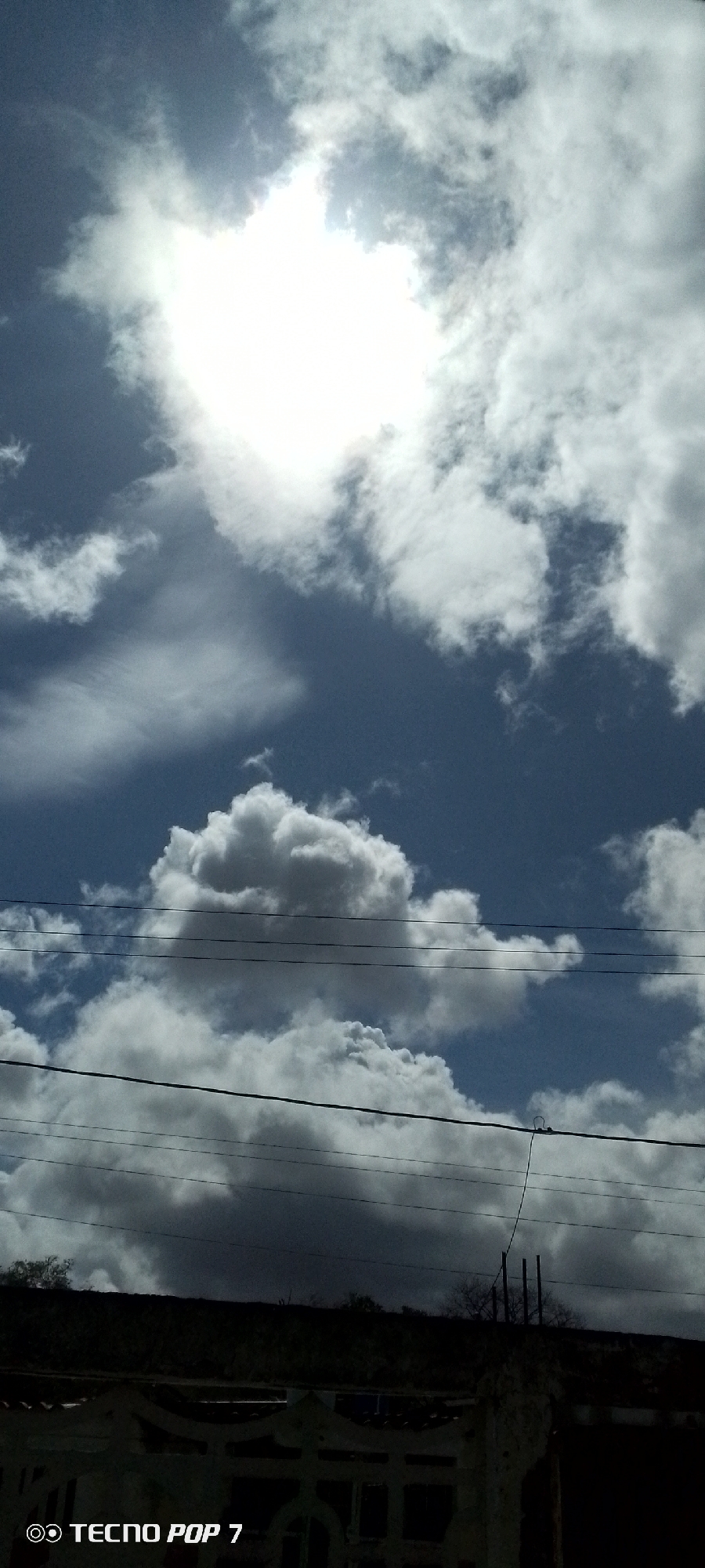 The image shows a bright sky with scattered white and gray clouds, obscuring a portion of the sun, which appears as a bright white glow at the top center. Below the sky, there are multiple horizontal power lines stretching across the frame. At the bottom of the image, a dark silhouette of a building or structure is visible, with some details of a fence or wall in the foreground. The lower left corner of the image contains a watermark that reads "TECNO POP 7". The scene is likely an outdoor view from the city of Altagracia, Venezuela, possibly depicting a residential or urban area with overhead electrical infrastructure.
