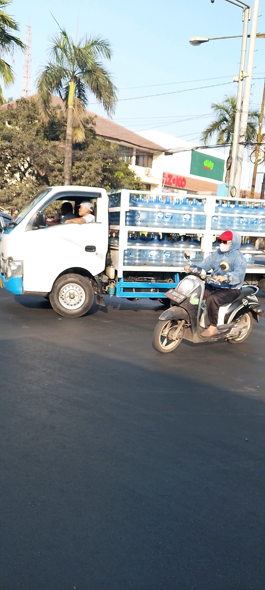 In Bekasi, Indonesia, a white truck carrying large blue water bottles is traveling on a paved road. Two men are visible in the truck's cabin, the driver and a passenger, both wearing white hats. A person on a dark-colored scooter is riding alongside the truck, wearing a blue hooded jacket, a red cap, sunglasses, and a white mask. The scooter has a license plate that reads "D 3530 TB 09-27". In the background, palm trees, buildings with brown roofs, and a green sign with red lettering are visible. The sky is clear and blue.