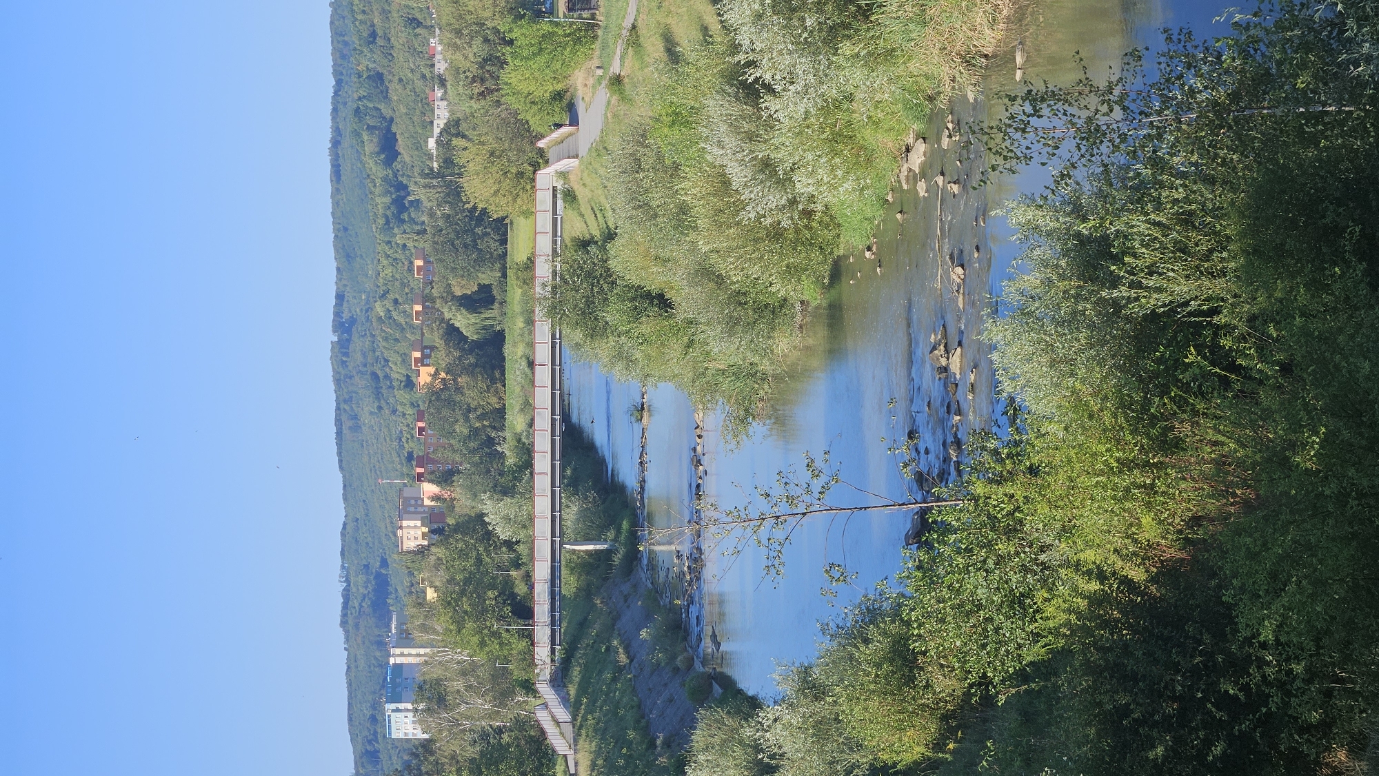 The image shows a sunny day with a clear blue sky over a city and a river. A pedestrian bridge spans across a moderately wide river, with its banks lined with lush green vegetation. The bridge is white with red railings. On the far bank, a pathway leads away from the bridge. In the background, apartment buildings and trees are visible on a gently rolling landscape. The river itself appears shallow with rocks visible beneath the surface. The scene is peaceful and natural, with a touch of urban development in the distance. This is likely the Sekčov river in Prešov, Slovakia, given the distinctive apartment buildings visible in the background which are characteristic of the Sekčov housing estate.